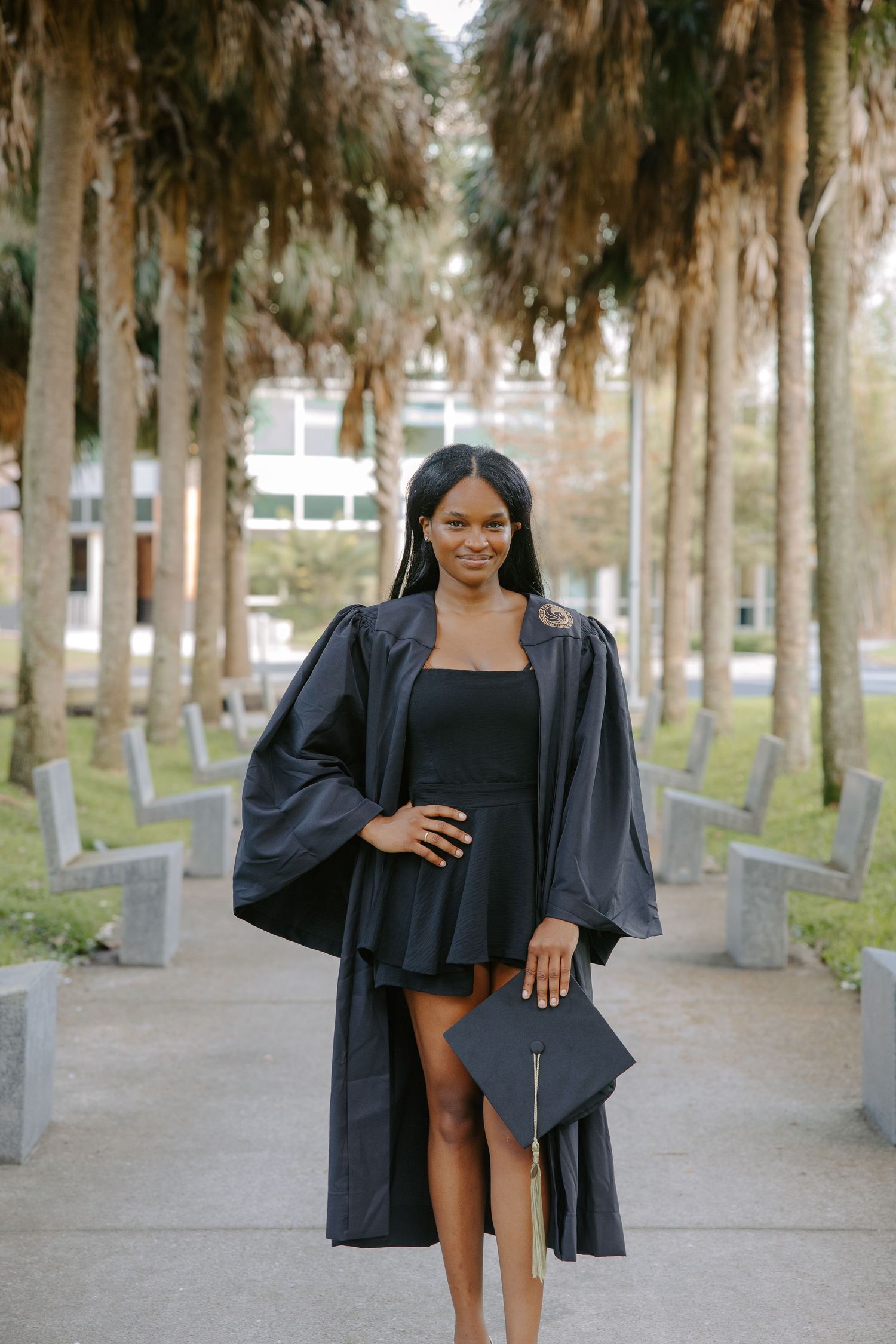 Graduate in black graduation gown stands elegantly among palm trees on campus pathway.