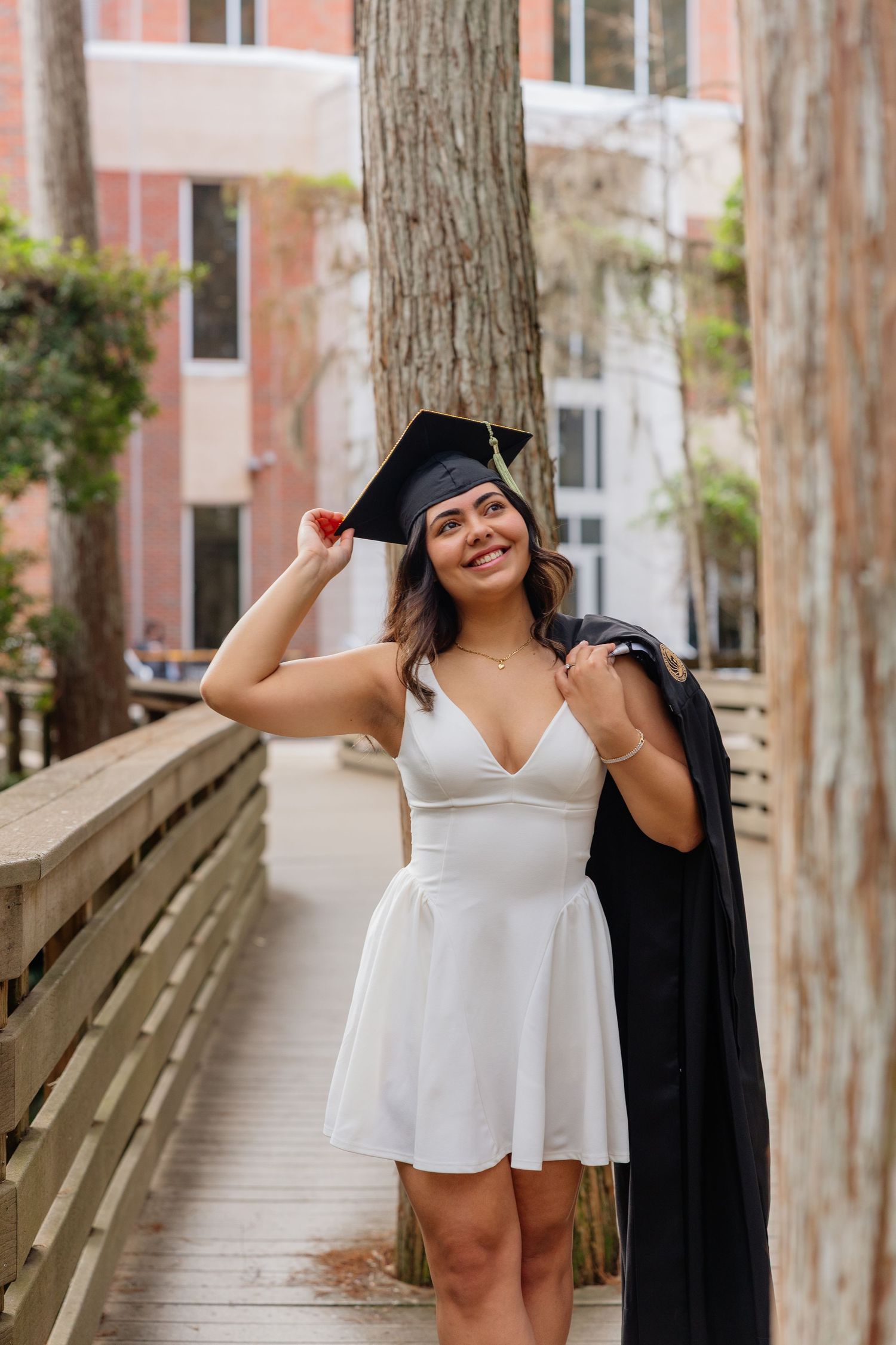 A graduate in cap and gown poses joyfully on a wooden walkway surrounded by trees and buildings.