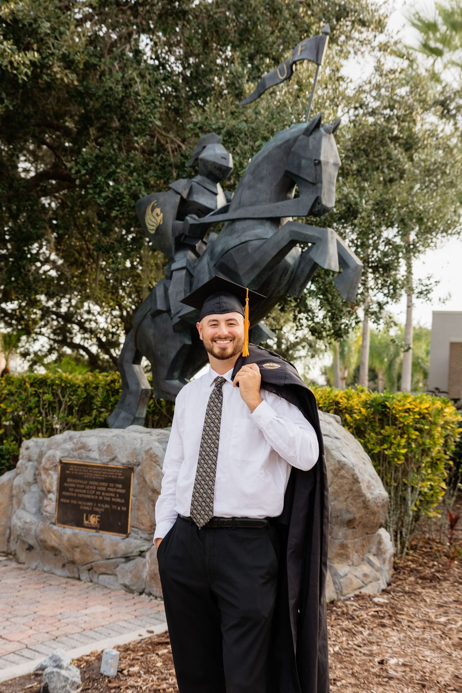 A graduate poses in front of a stone monument wearing a black graduation gown and cap.