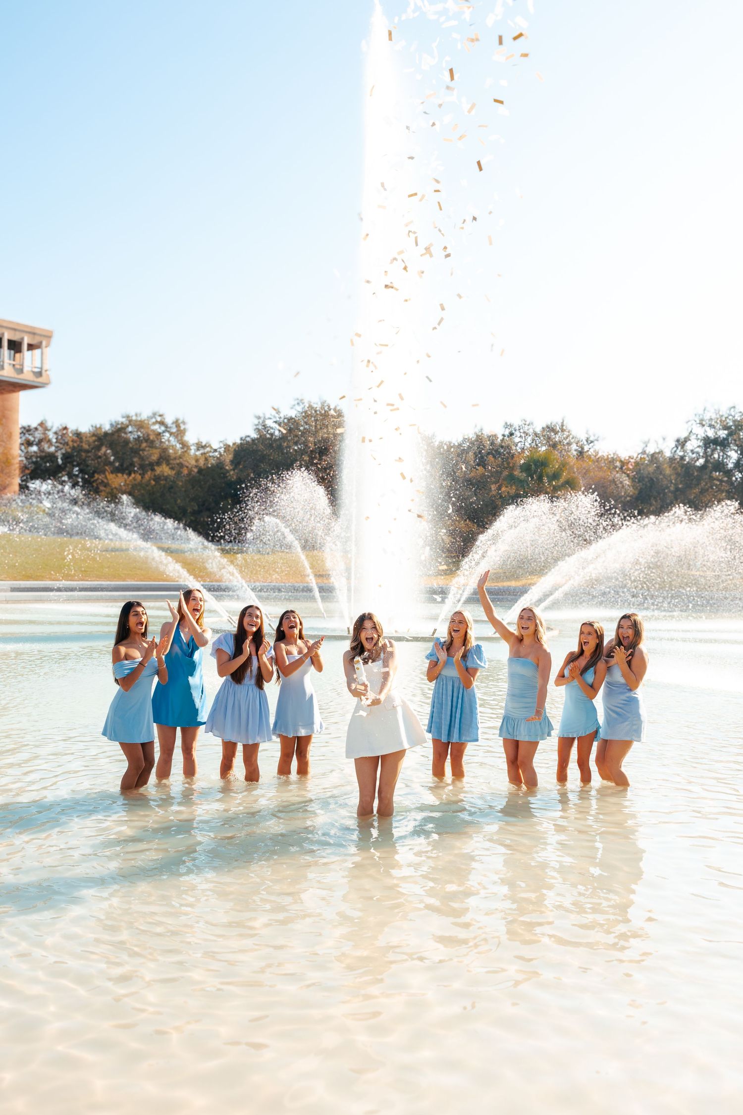 Group wearing matching blue dresses splashes and plays in shallow beach water during sunset photoshoot.