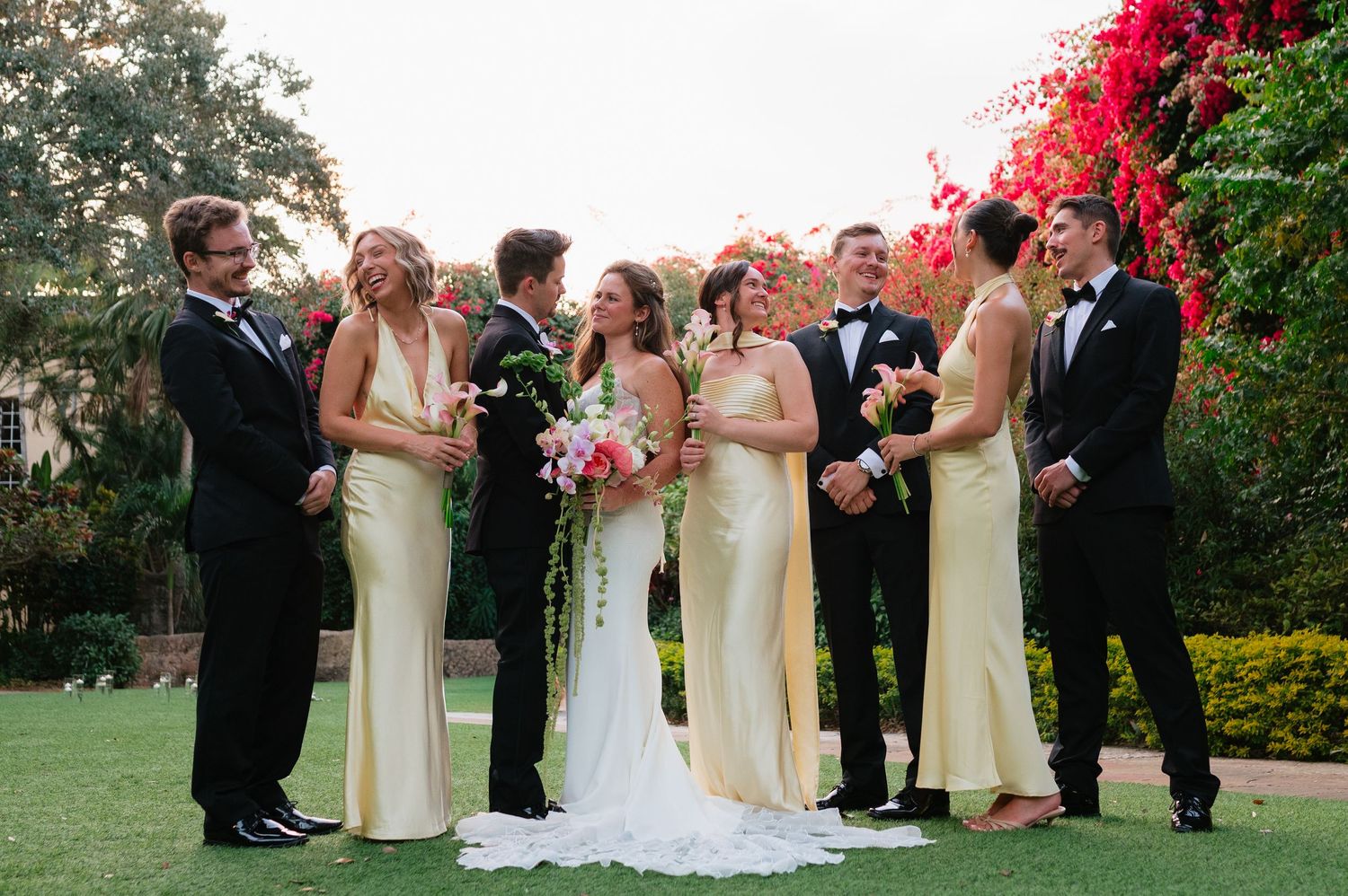 Wedding party in formal attire poses on lawn with vibrant red bougainvillea flowers in the background.