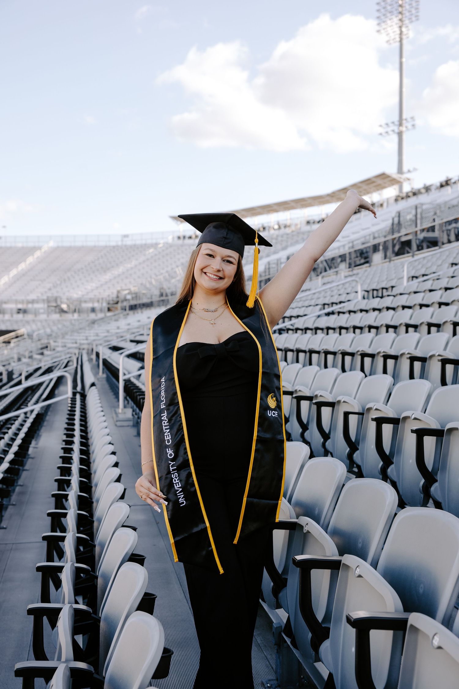 Graduate in black cap and gown poses in empty stadium seats.