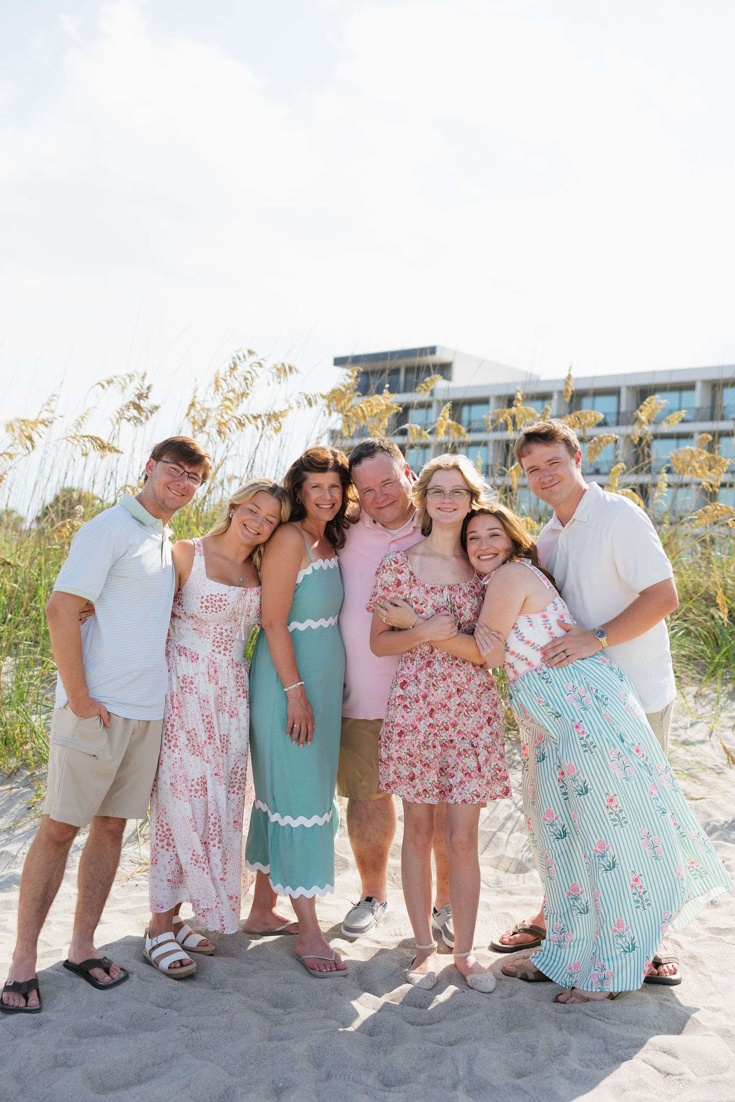 Happy family of seven posing together on a sandy beach, wearing coordinated pastel outfits with sea oats in the background.