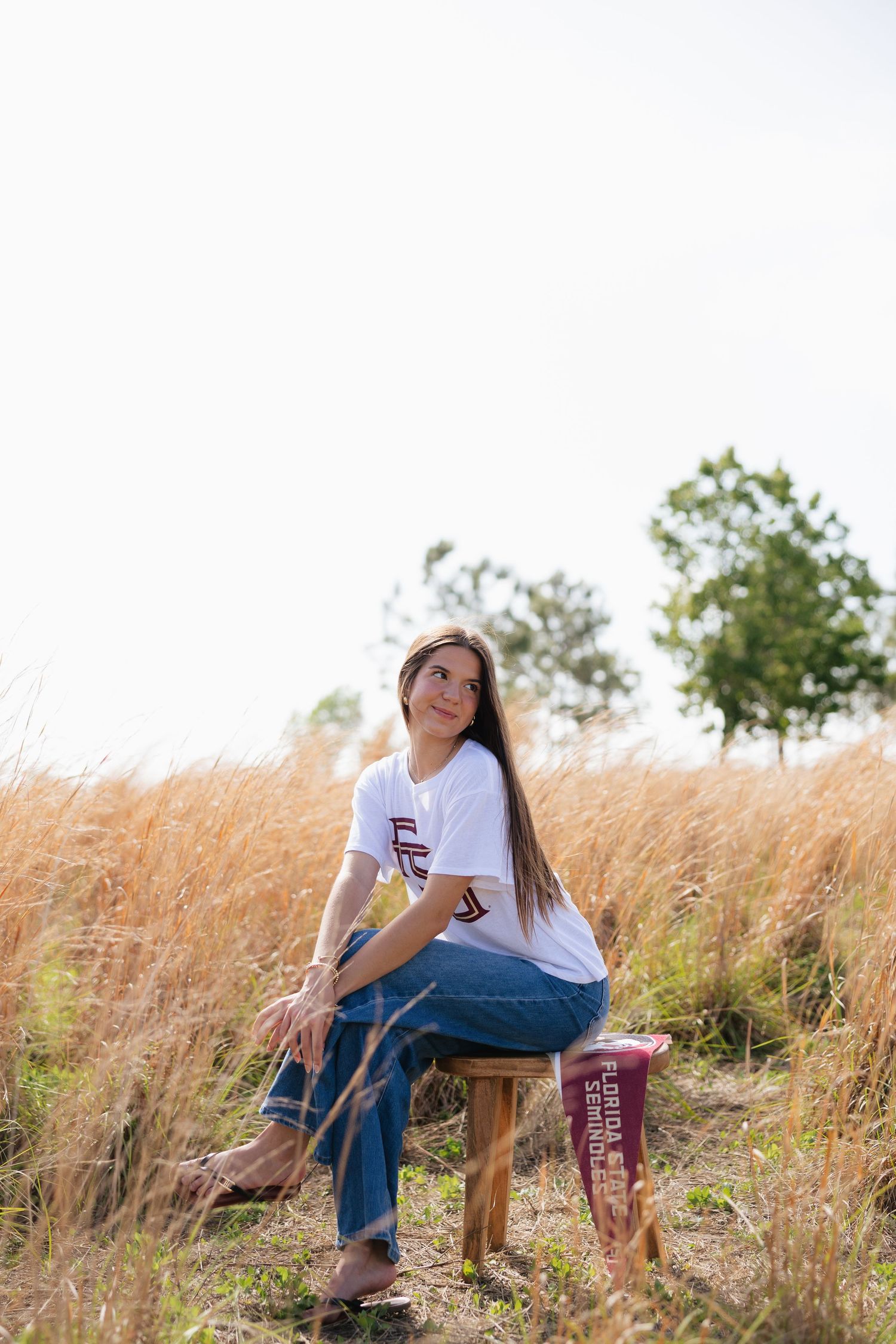 Young woman in FSU shirt and jeans sits on wooden stool in golden field, gazing upward on sunny day.