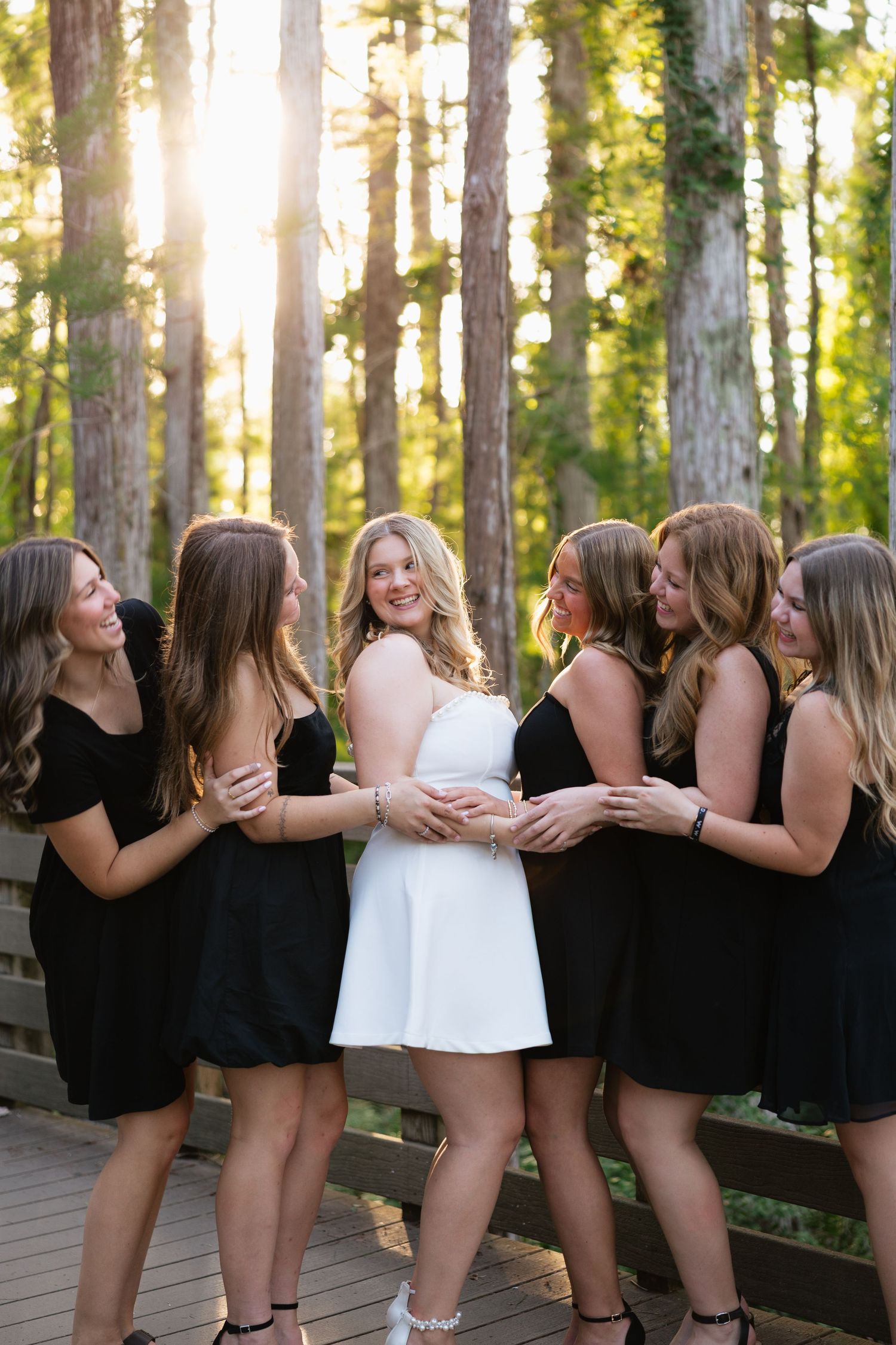 Bride in white dress surrounded by bridesmaids in black dresses, posing on a wooden deck in a sunlit forest.