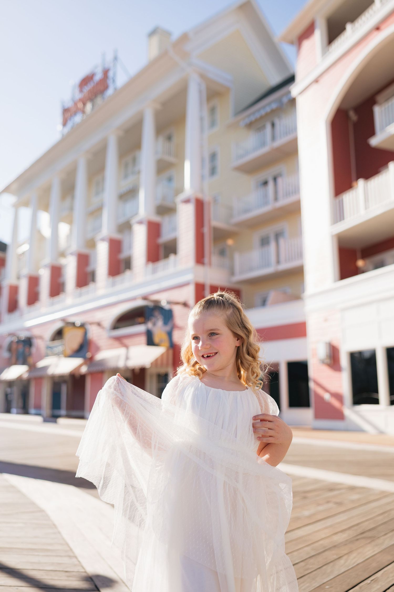 Young girl in white dress smiling on sunny boardwalk with pink and white hotel building in background.