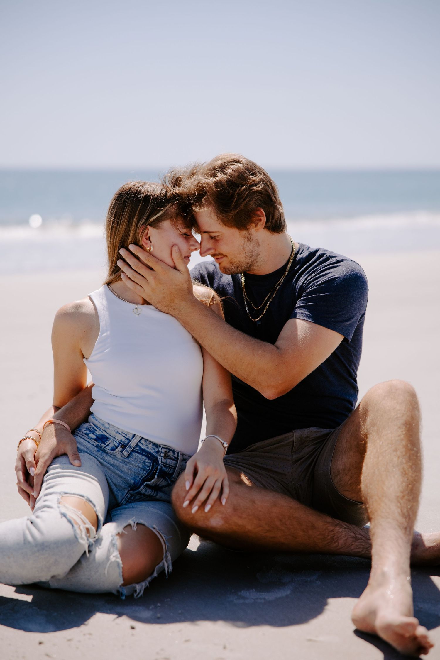 A couple sits close together on a sandy beach wearing casual summer clothes as they share an intimate moment.