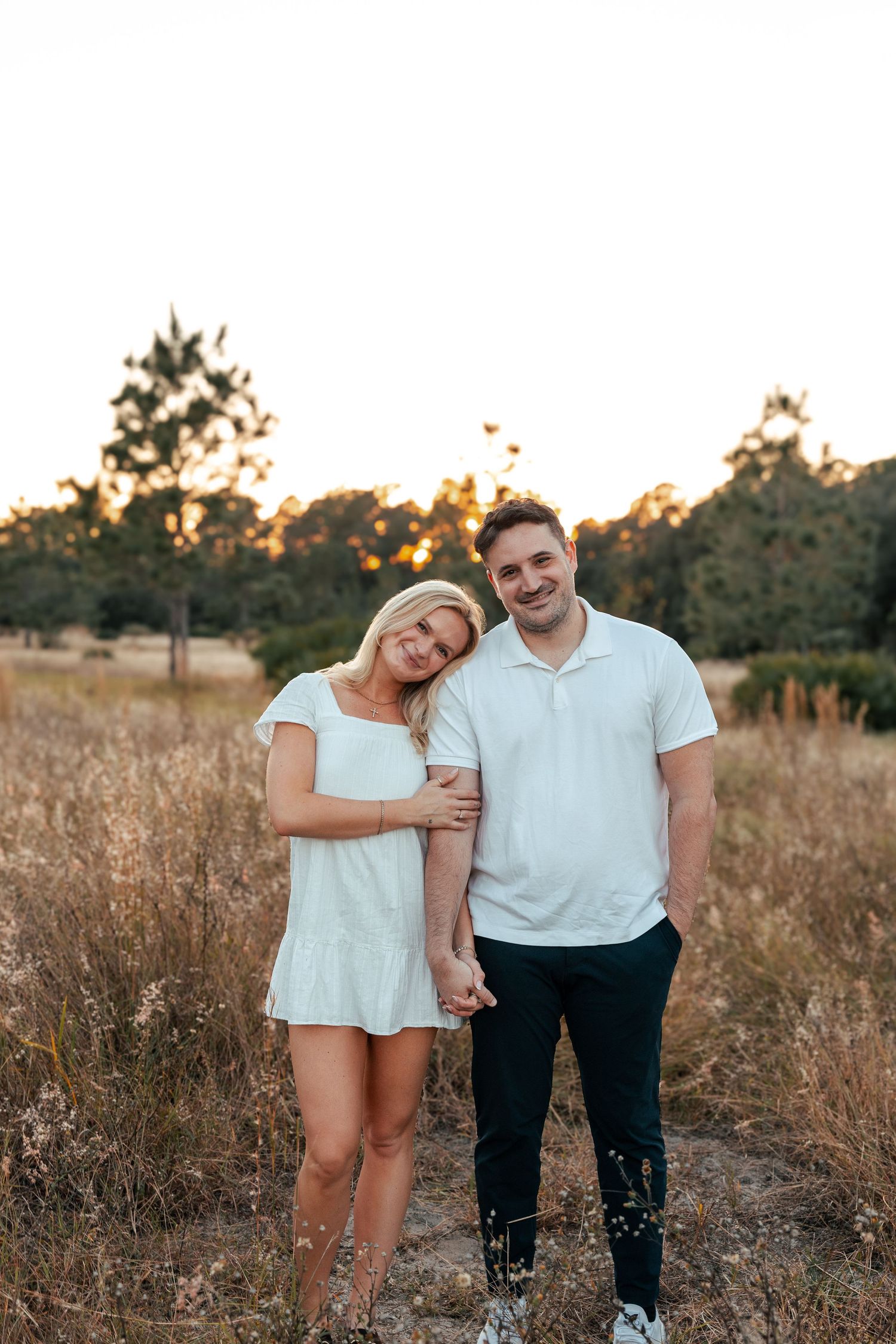 A couple stands together in a golden field during sunset, captured in a series of romantic outdoor portraits.