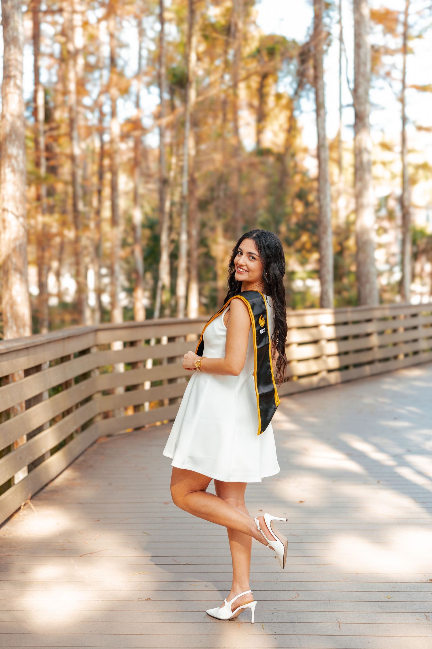 A woman in a white summer dress poses on a wooden boardwalk surrounded by autumn trees in golden sunlight.
