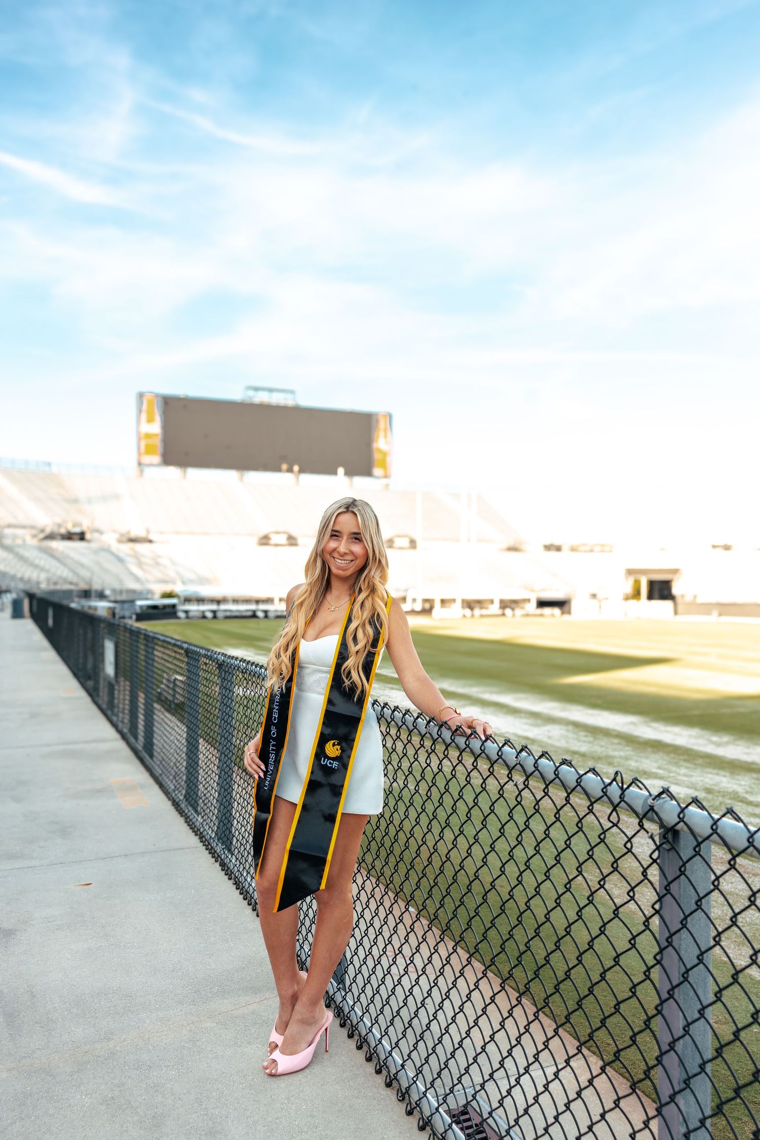 A graduation photoshoot featuring someone in academic regalia posing by a chain link fence at a sports field.