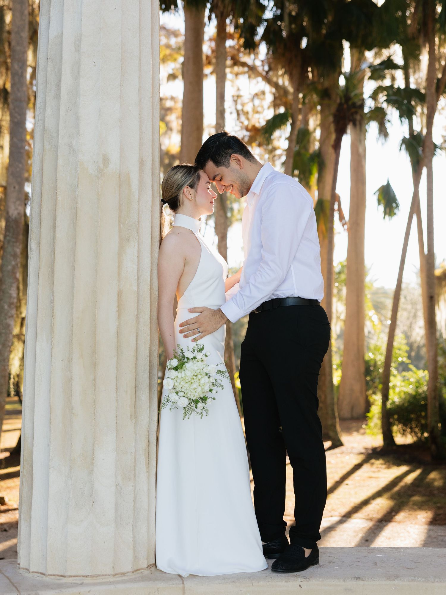 A romantic wedding couple shares an intimate moment by a white column in a sunlit eucalyptus grove.