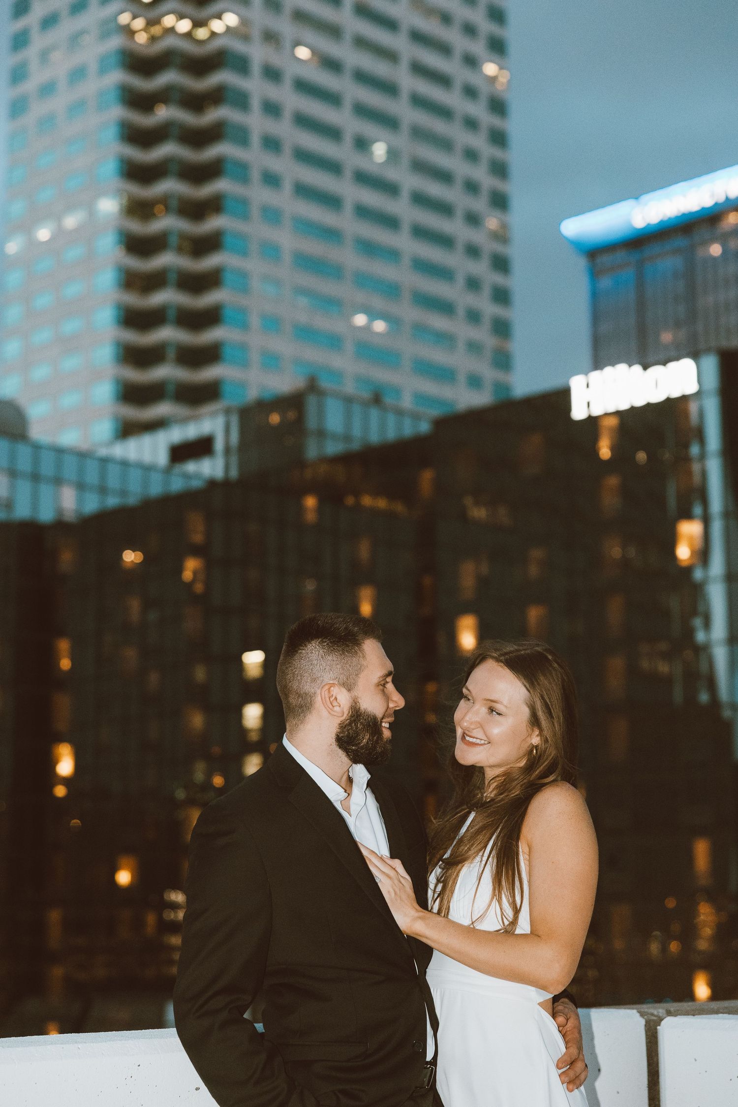 A couple shares an intimate moment on a rooftop terrace with city lights and high-rise buildings in the background.
