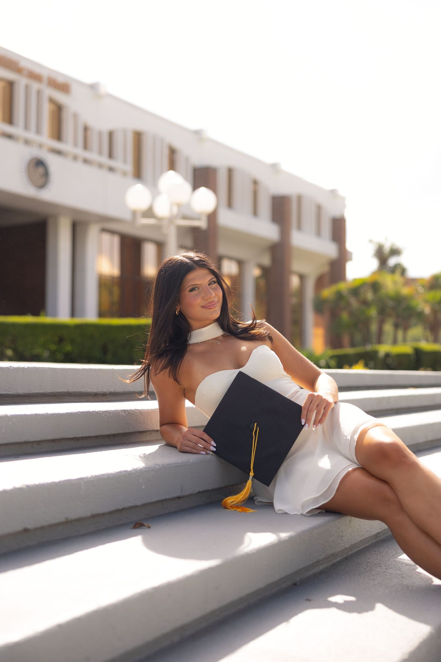 Young woman in white dress holding graduation cap sits on sunlit steps outside university building.