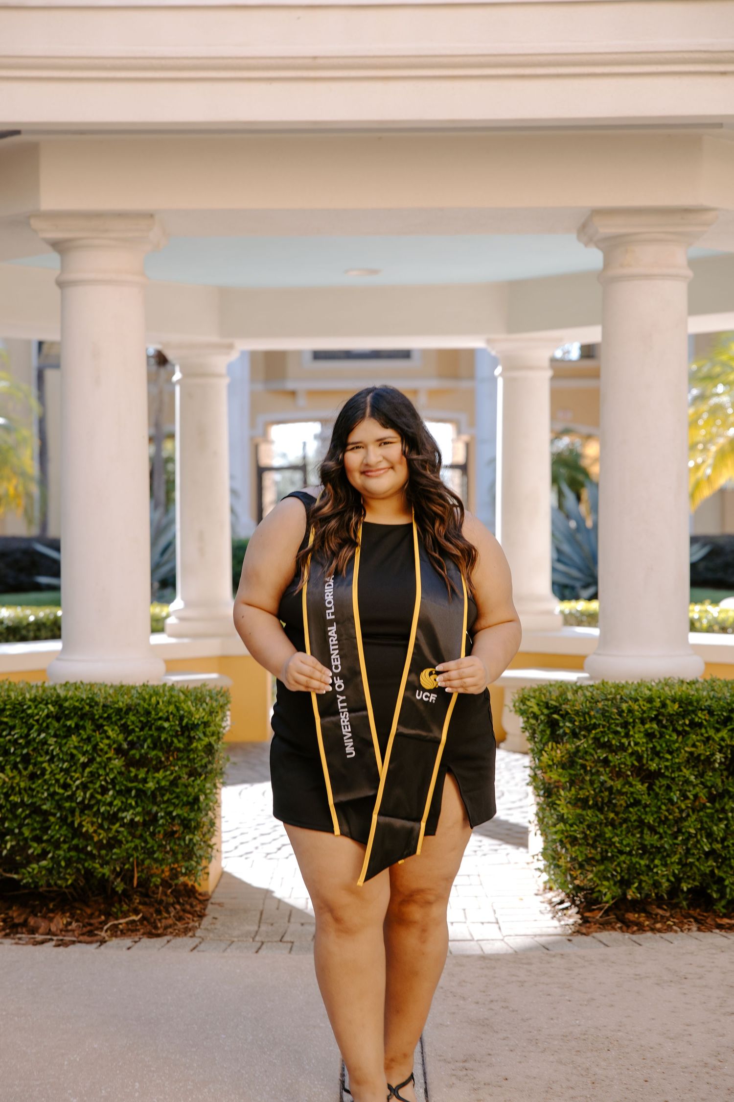 Graduate in black dress with gold sash poses in front of columned building.