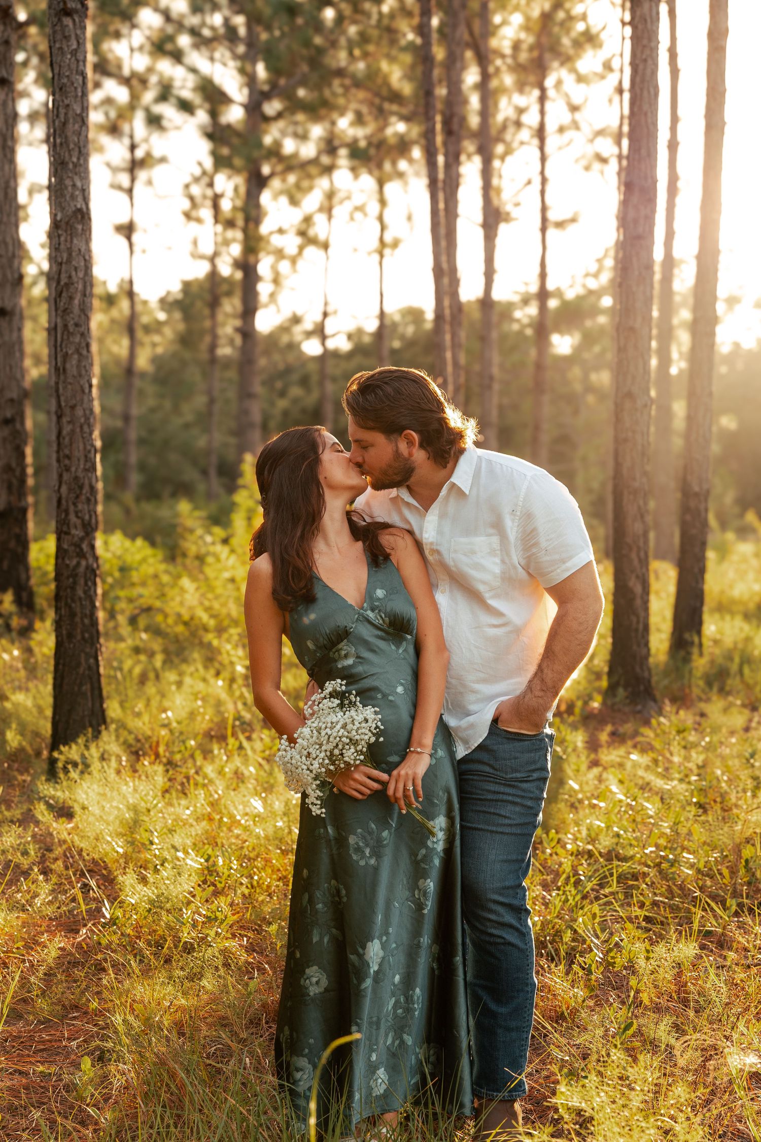 Romantic couple sharing a tender moment in a sunlit forest while holding wildflowers at golden hour.