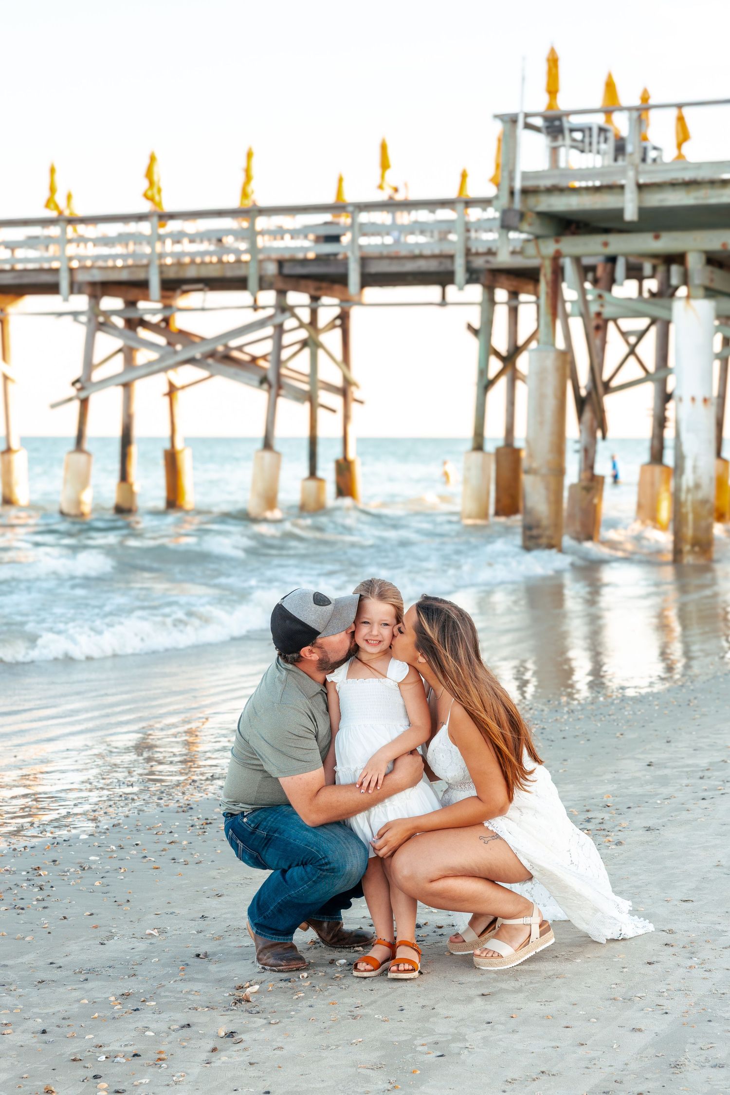 A couple shares tender moments on a sandy beach near a wooden pier at sunset.