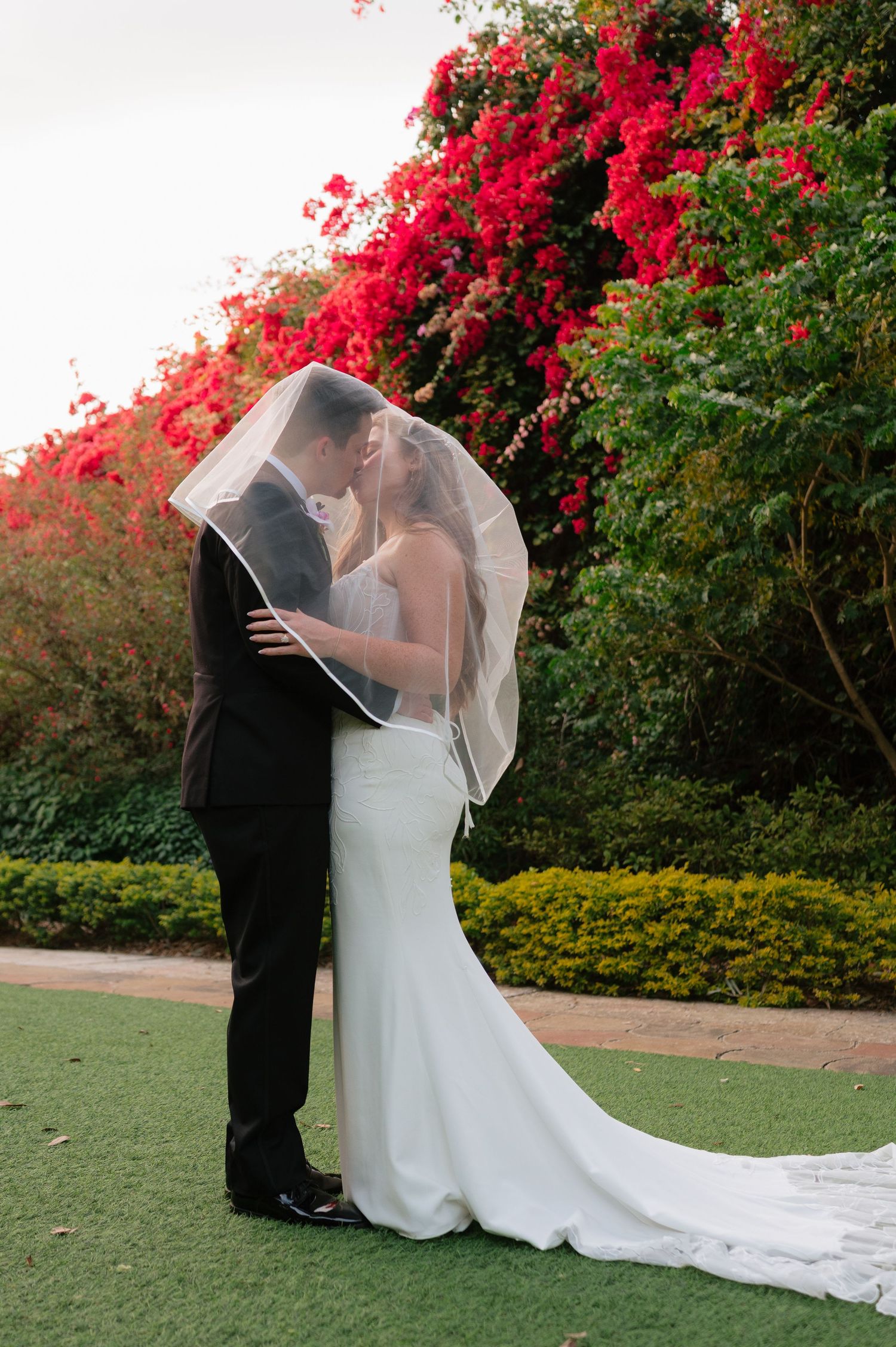 A romantic wedding couple embraces under vibrant red bougainvillea flowers in a garden setting.