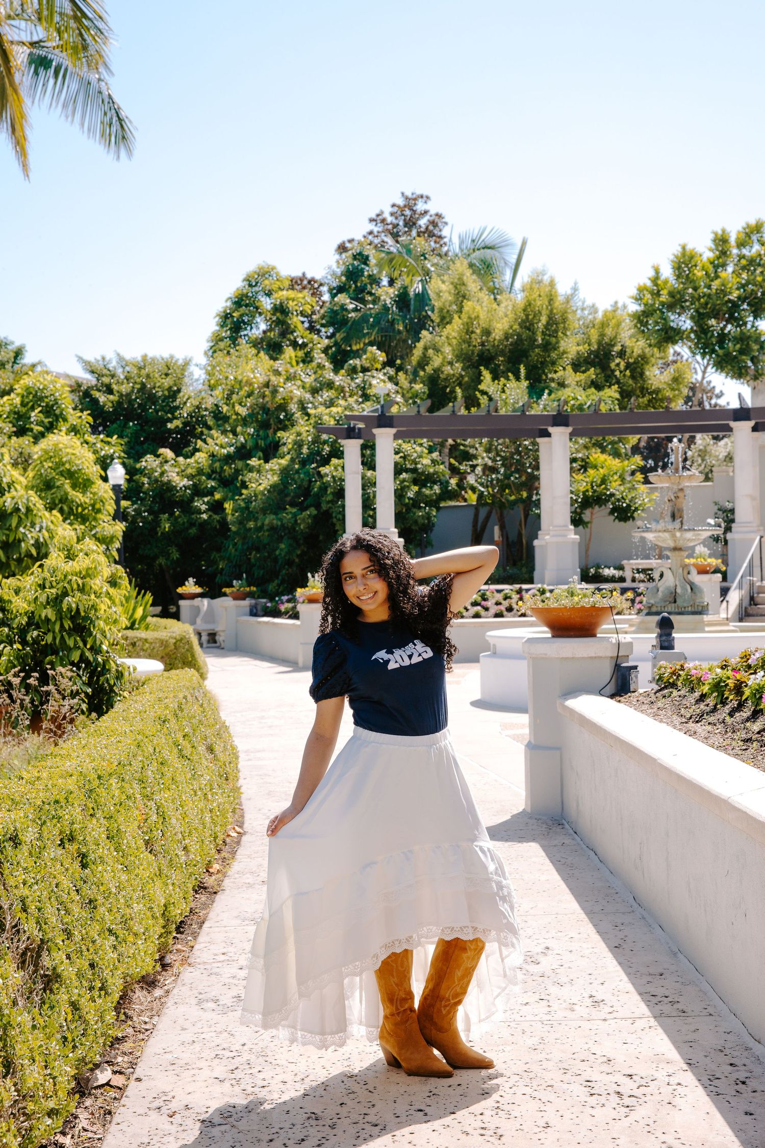 Graduate in black top and white skirt poses in garden setting with pergola.
