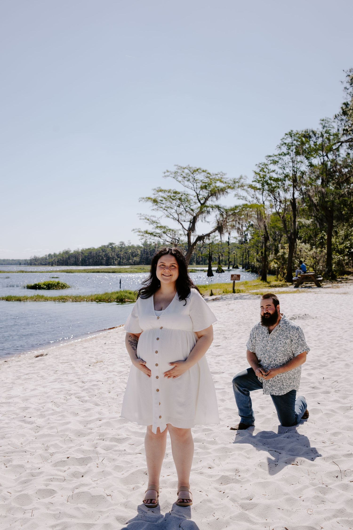 A scenic beach photoshoot shows people posing and having fun on white sand with trees and water in the background.
