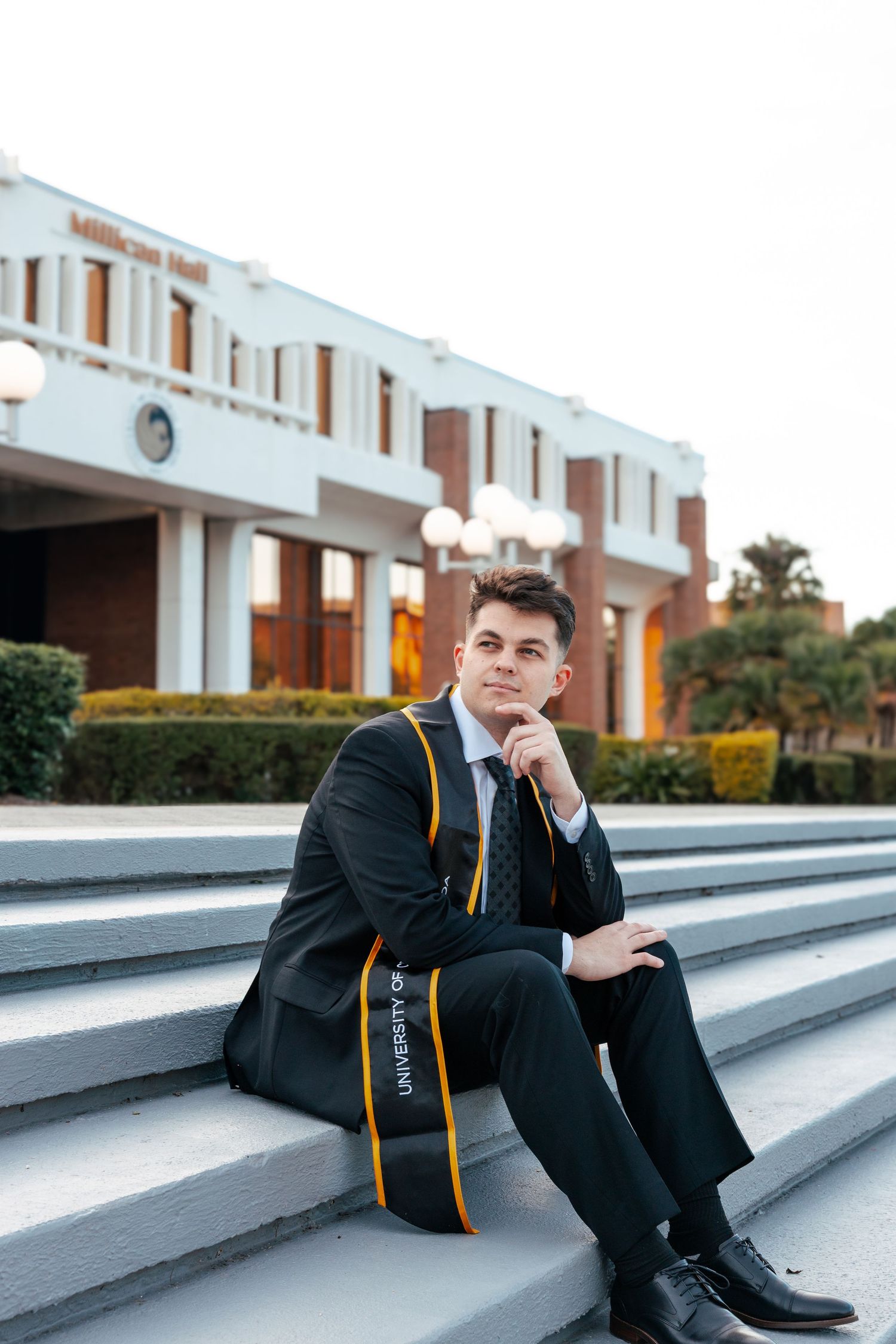 A person in a black suit with gold stripes sits on concrete steps outside a modern building.