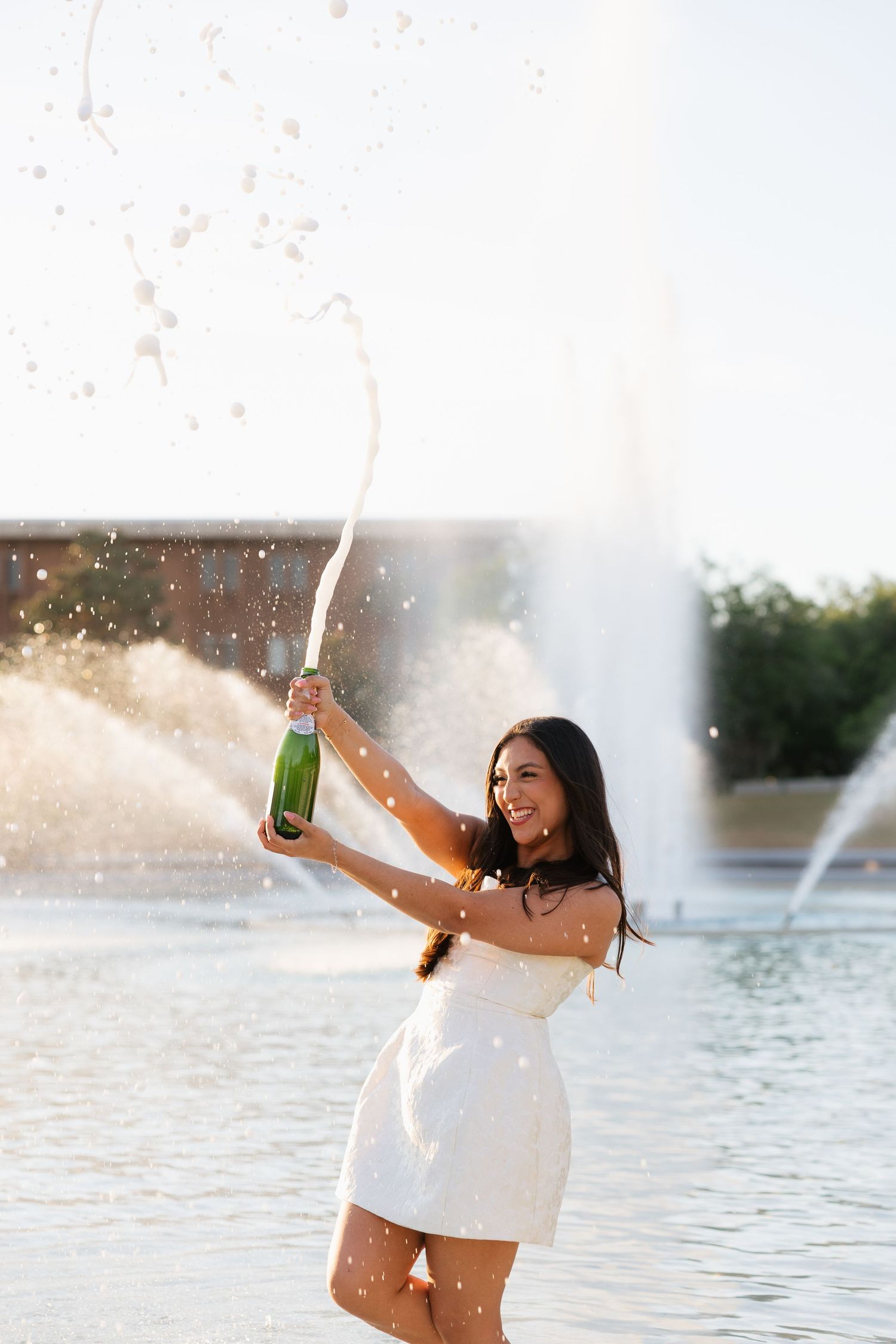 Young woman in white dress joyfully spraying champagne from bottle outdoors near fountain on sunny day.