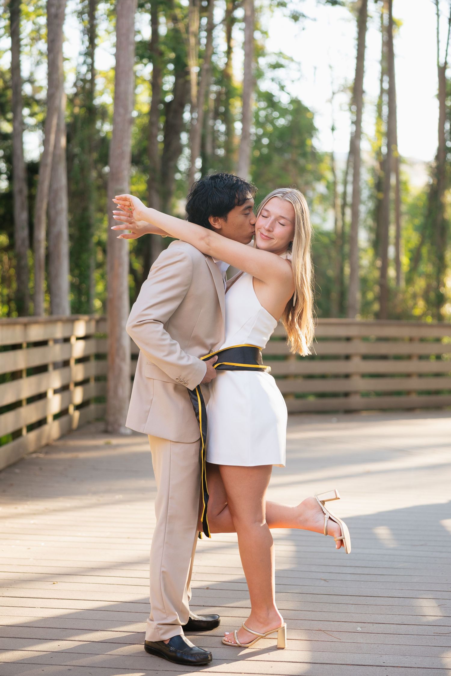 Couple sharing a romantic kiss on a wooden bridge, man in tan suit, woman in white mini dress, forest backdrop.
