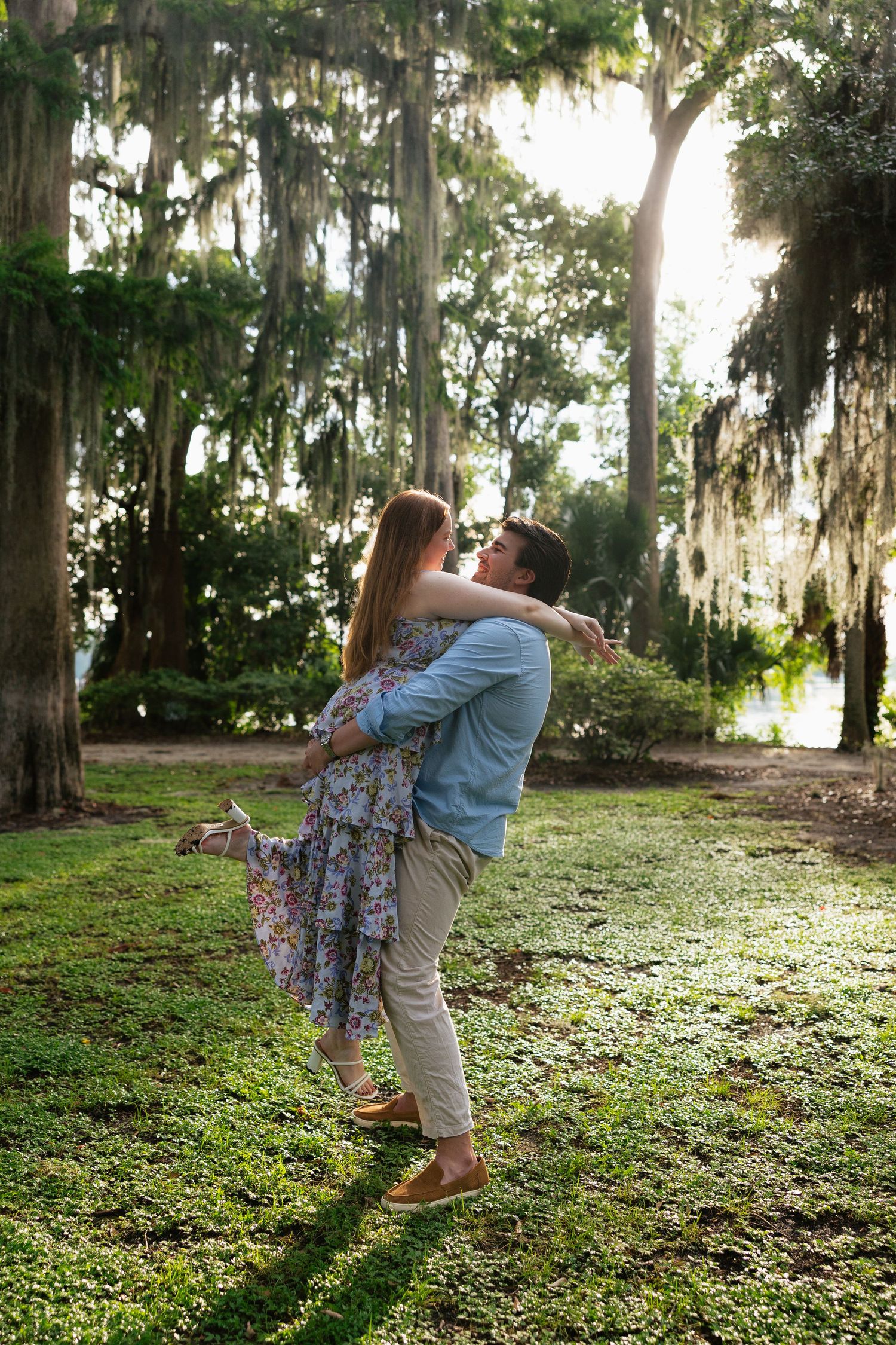 Couple embracing and spinning in a sunlit park with Spanish moss-draped trees and lush green grass.