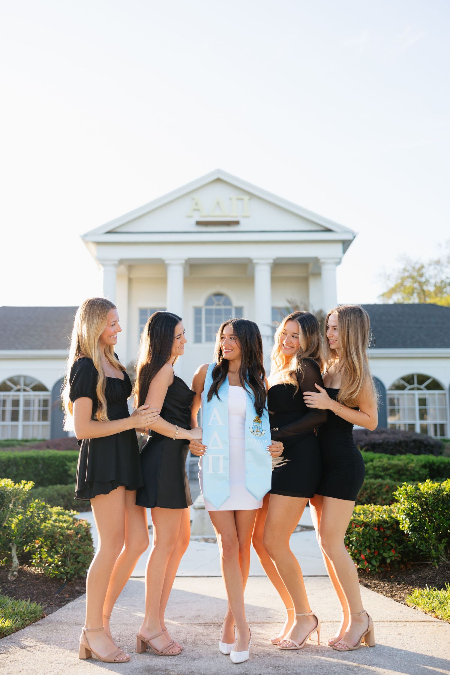 Five young women in black dresses pose together smiling outside a white colonial building on a sunny day.