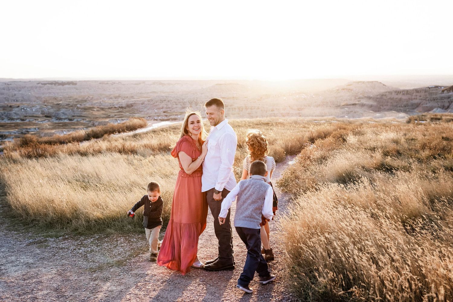 Family walking along a scenic hilltop trail through golden grass at sunset with mountains in the background.
