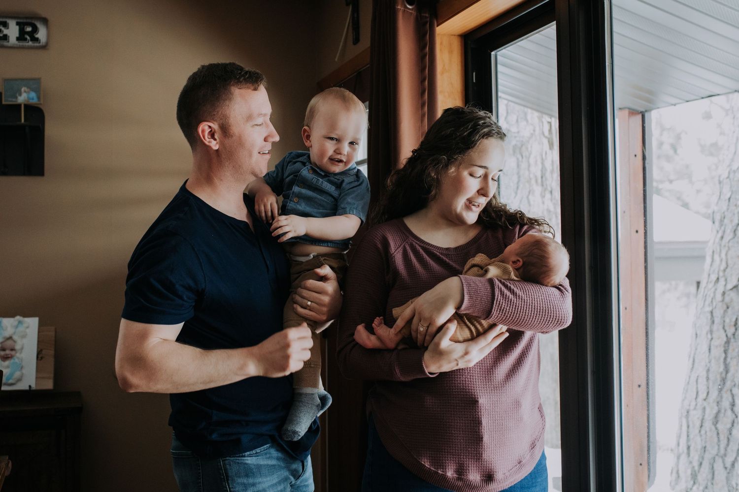 A family stands together by a window, two adults holding young children in their arms as they share a moment indoors.