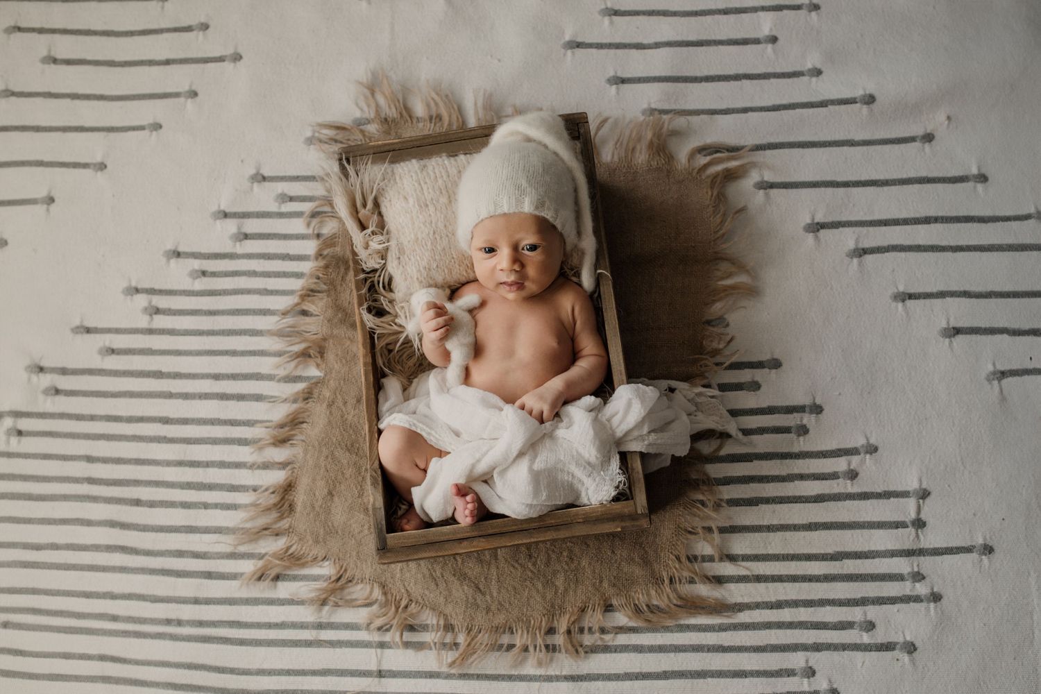A newborn baby wearing a knit bonnet lies peacefully on a fur blanket with white striped backdrop for a professional photo.