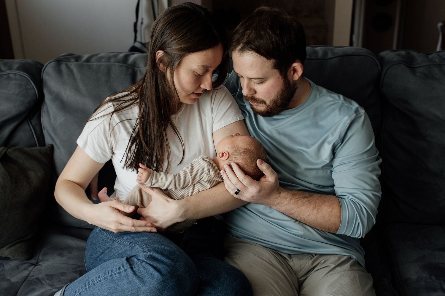 A couple sits together on a gray couch holding and gazing lovingly at their newborn baby.