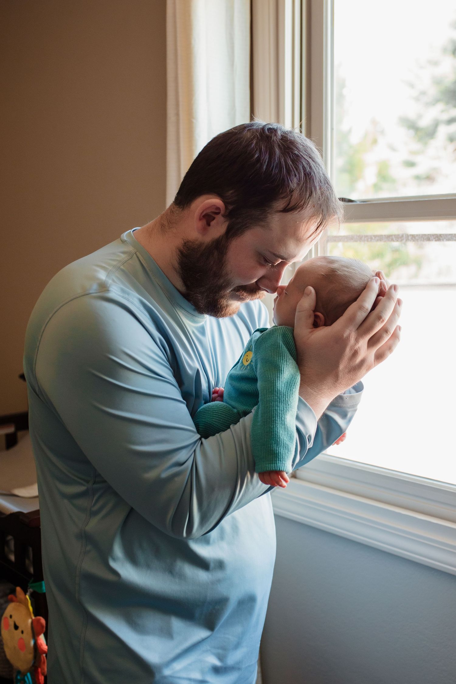 A tender moment by a window shows someone wearing a light blue shirt gently holding and kissing a small infant.