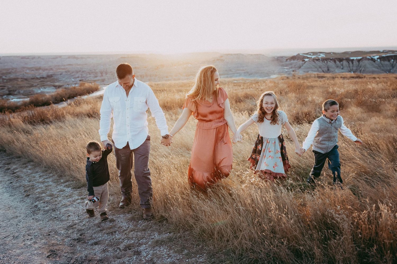 A family walks together holding hands through a golden grassy field at sunset.