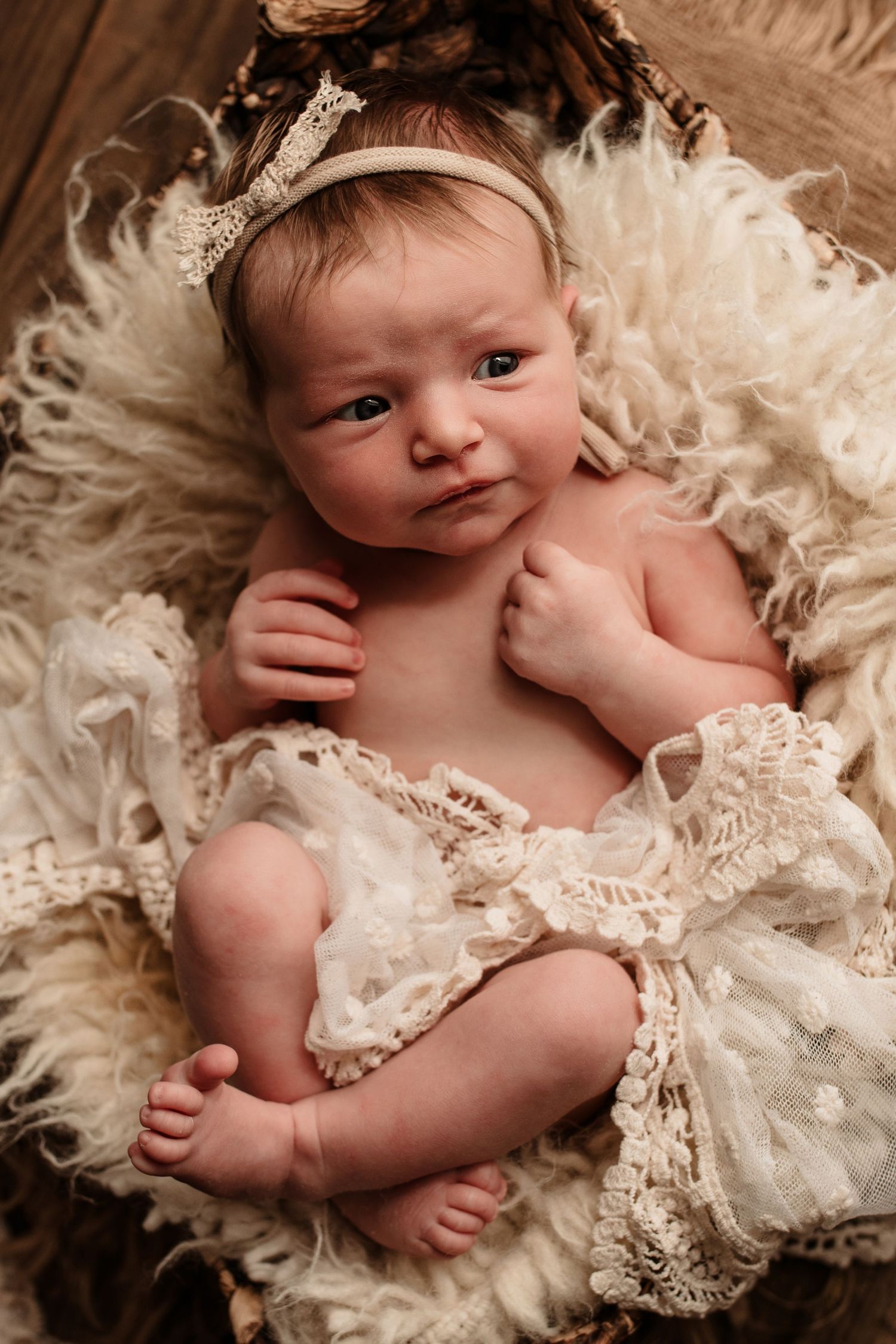 Sweet newborn baby lying on cream-colored lace and fur, wearing a sparkly headband while looking at camera.