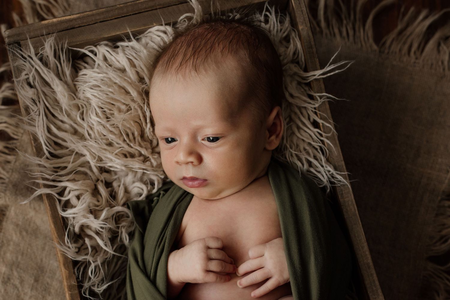 A newborn baby swaddled in olive green fabric rests on soft textured beige fur bedding.