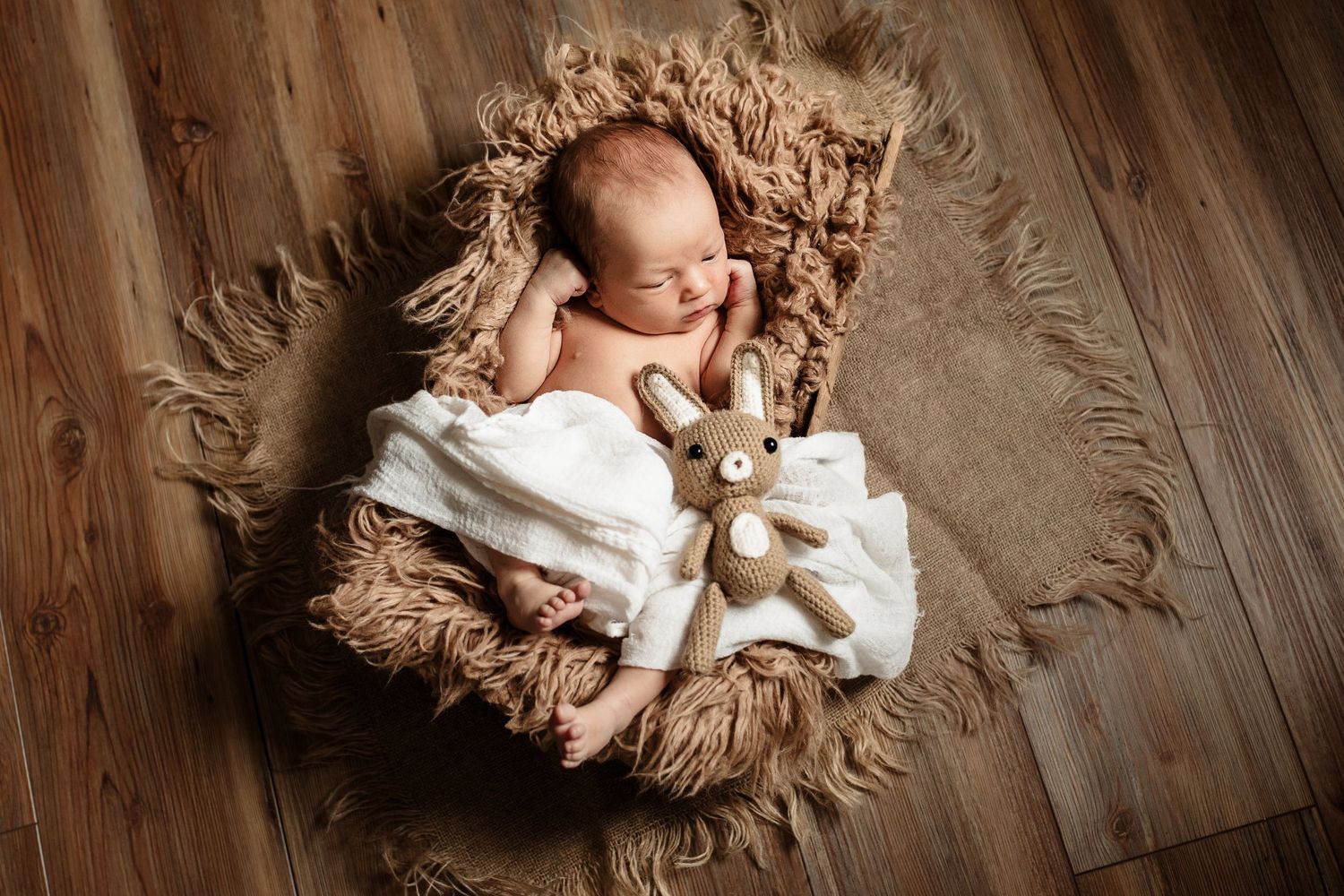 Newborn sleeping peacefully on fluffy brown fur blanket with small stuffed bear toy on wooden floor.
