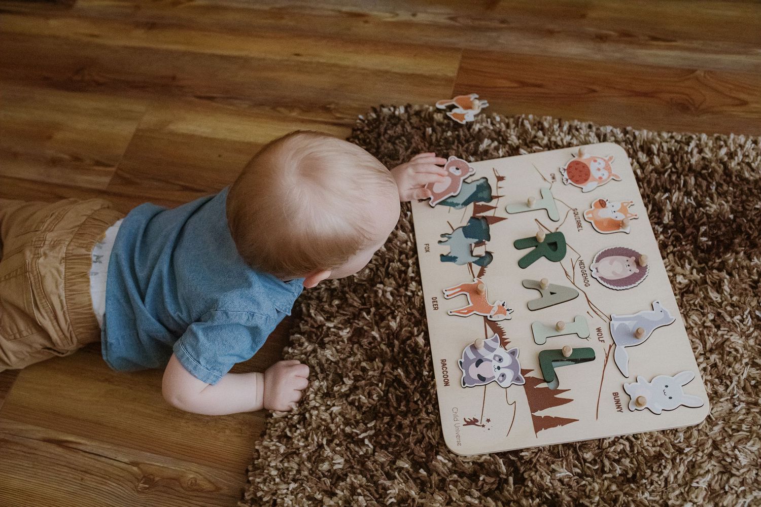 Baby in blue clothing lies on plush carpet while playing with an alphabet learning board on wooden floor.