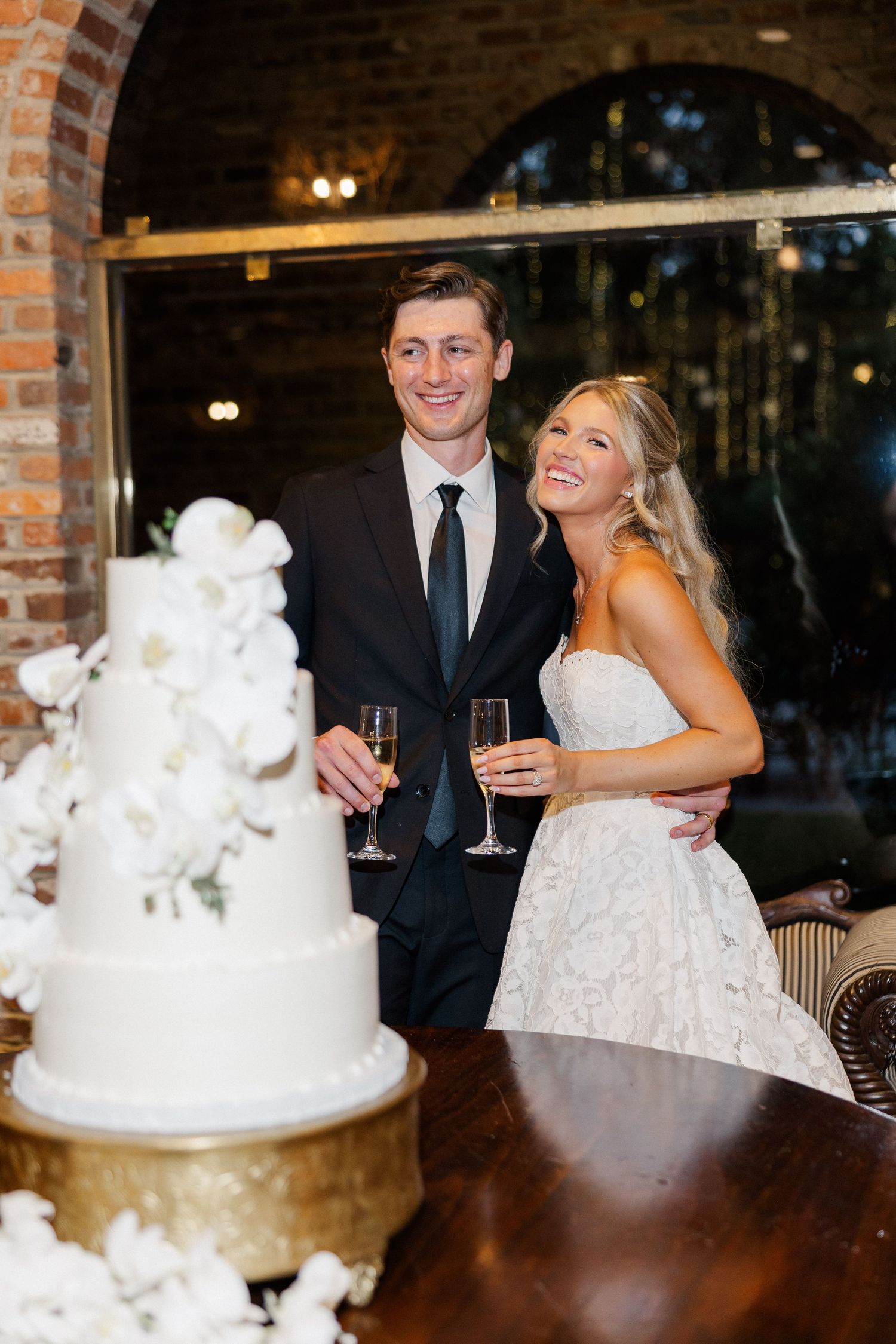 Smiling bride and groom toast with champagne beside elegant white floral wedding cake.