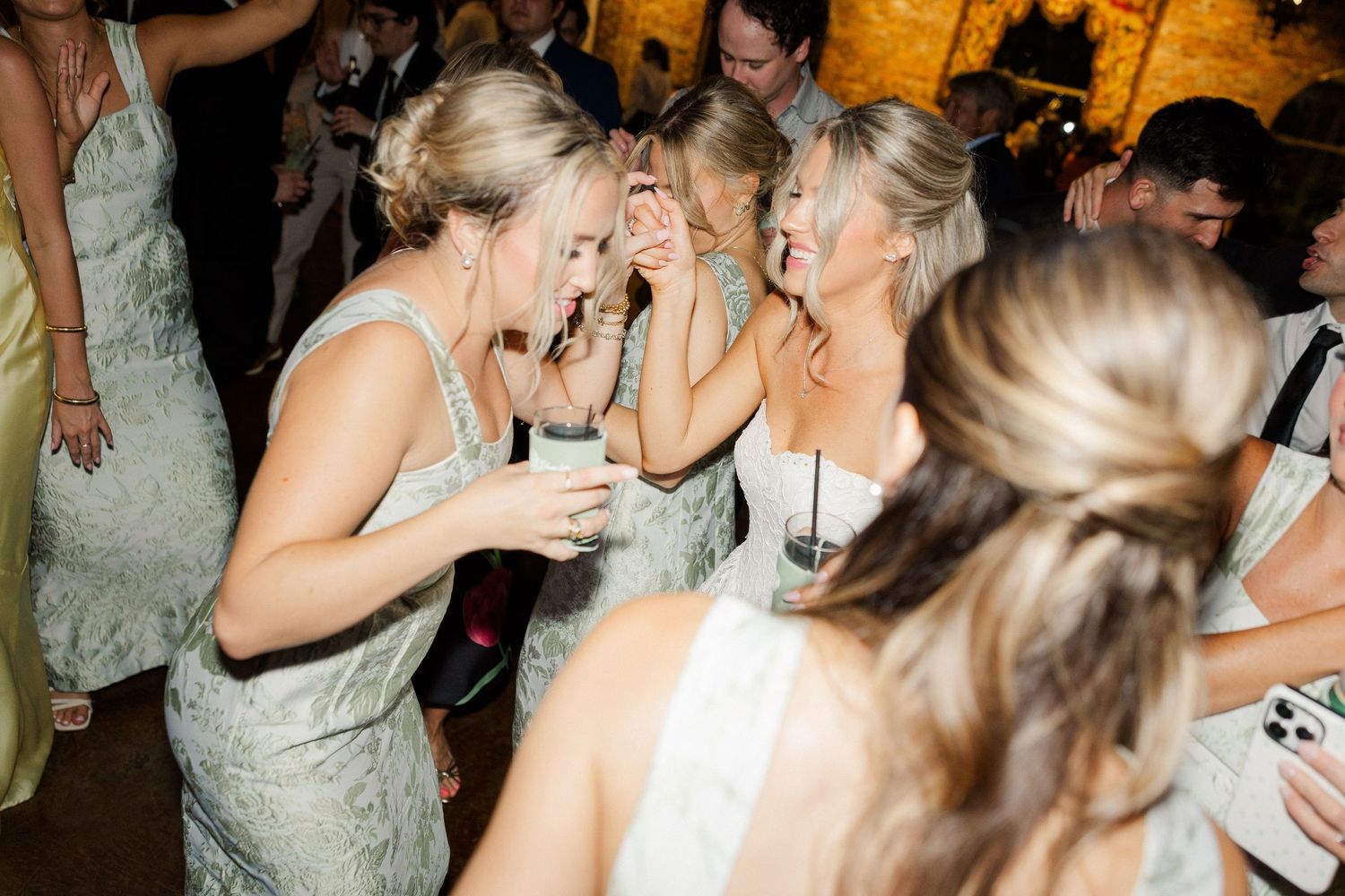 Bridesmaids in sage green dresses laughing and dancing together at wedding reception.