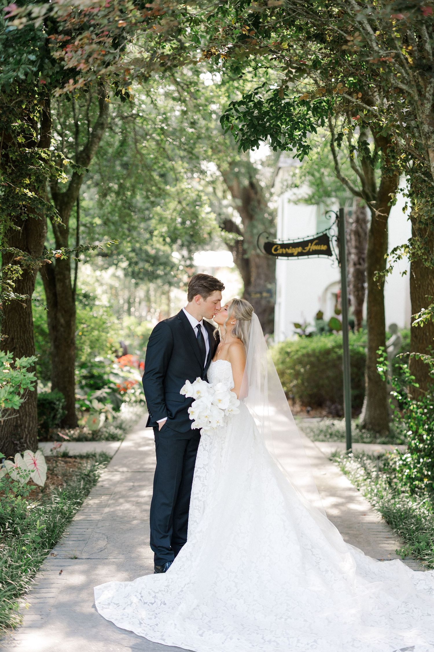 Bride and groom sharing a romantic kiss under a lush tree canopy near a Carriage House sign.