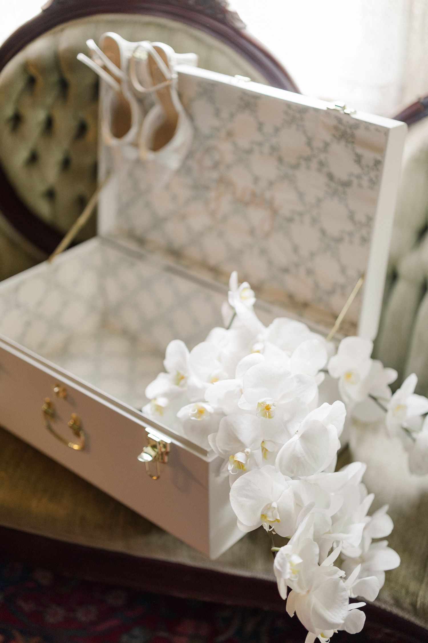 White orchids drape over an open decorative bridal box with wedding shoes reflected in a mirror.