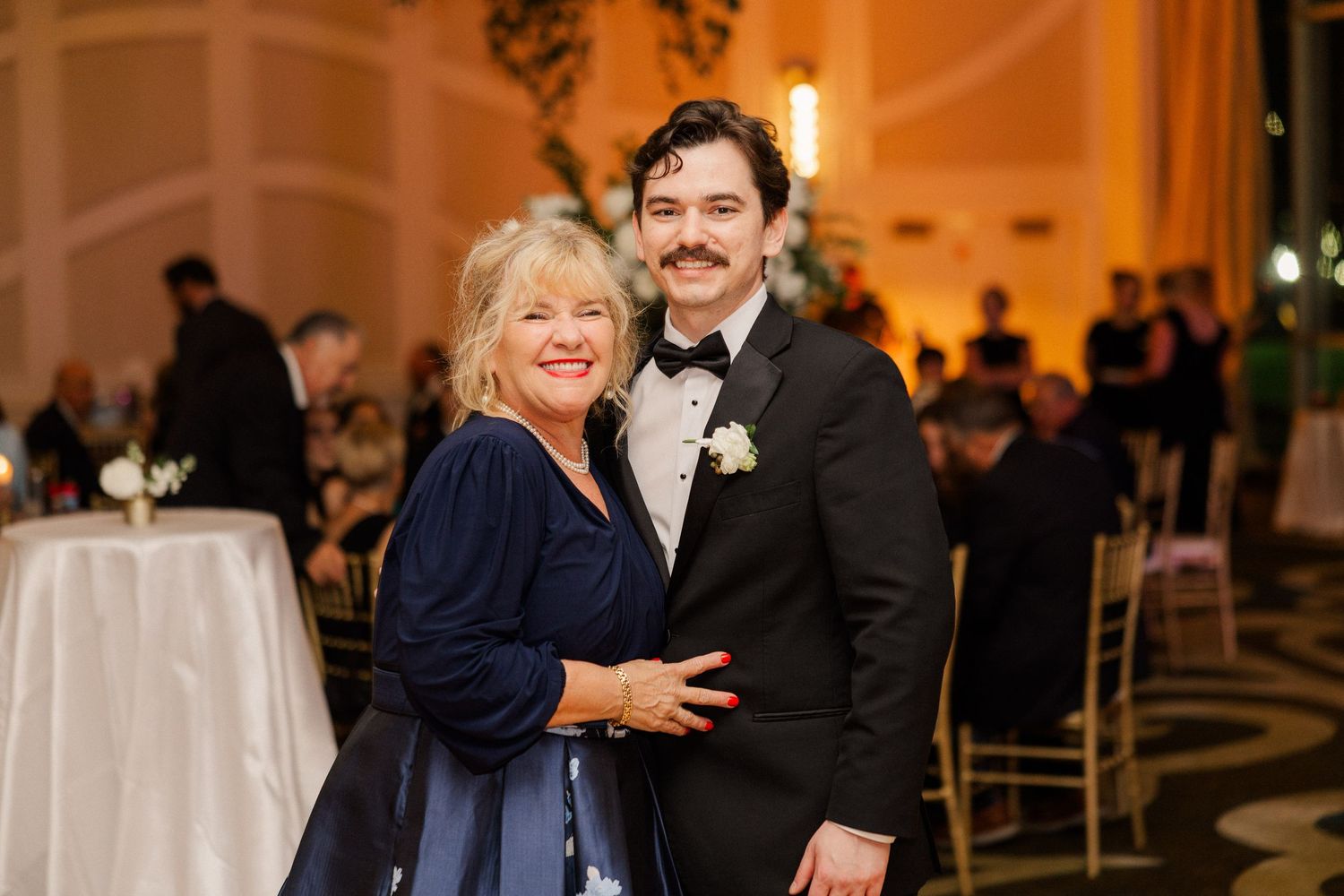 Groom with mustache in black tuxedo poses with smiling older woman in navy floral gown at wedding reception.