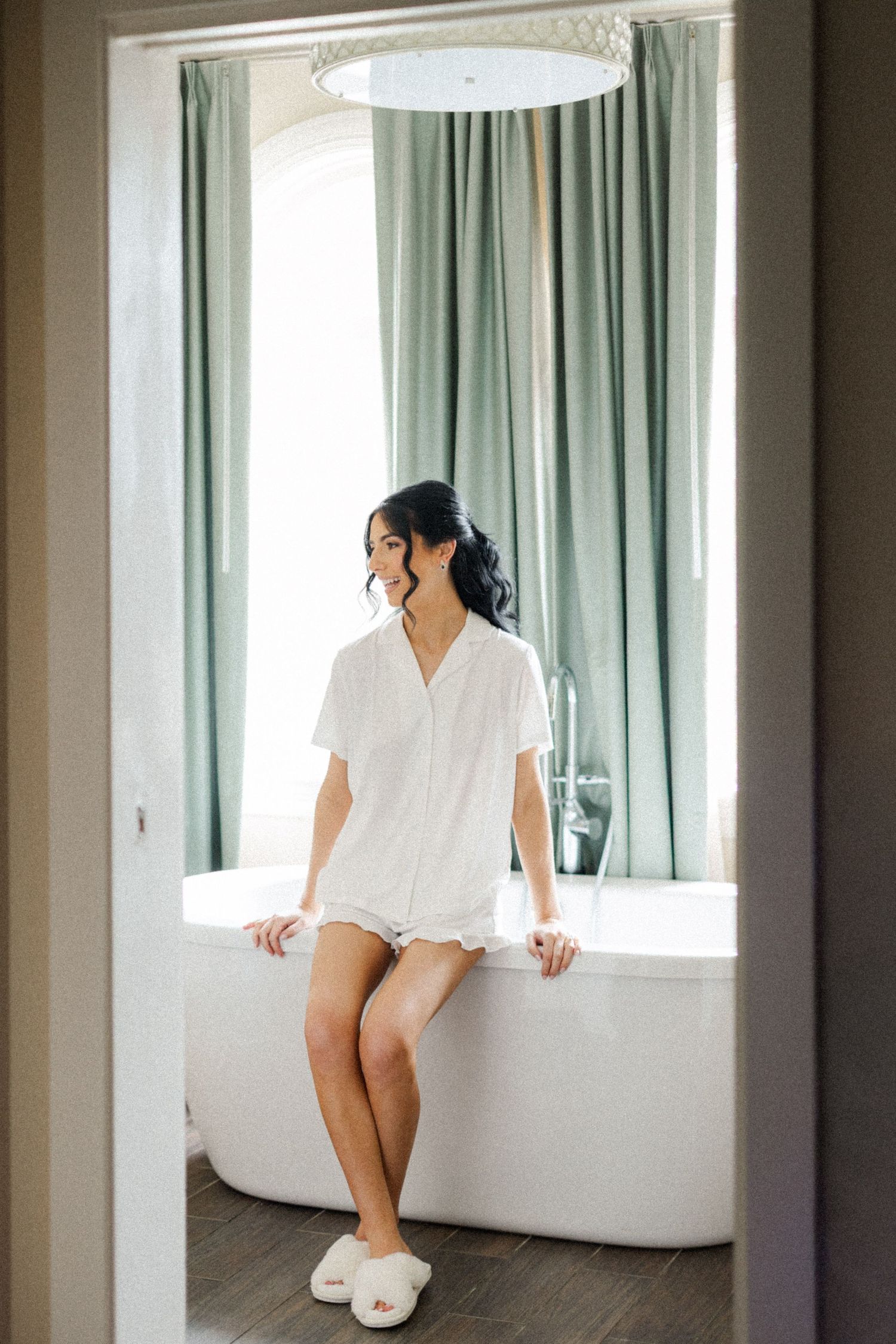 Bride in white robe sitting on freestanding bathtub in elegant bathroom with sage green curtains.