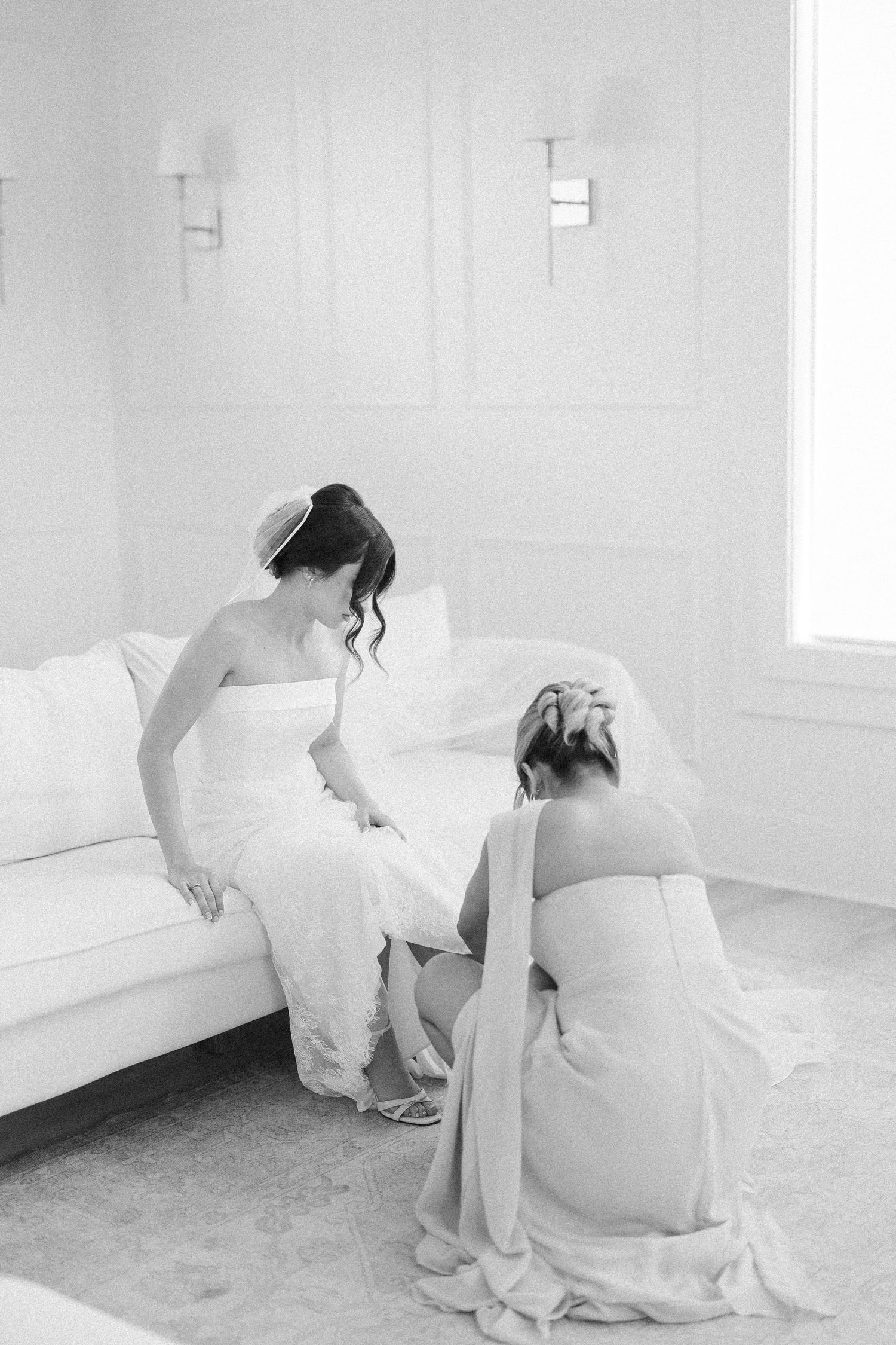 Black and white photo of a bride in a strapless gown seated on a sofa while a bridesmaid kneels to help her.