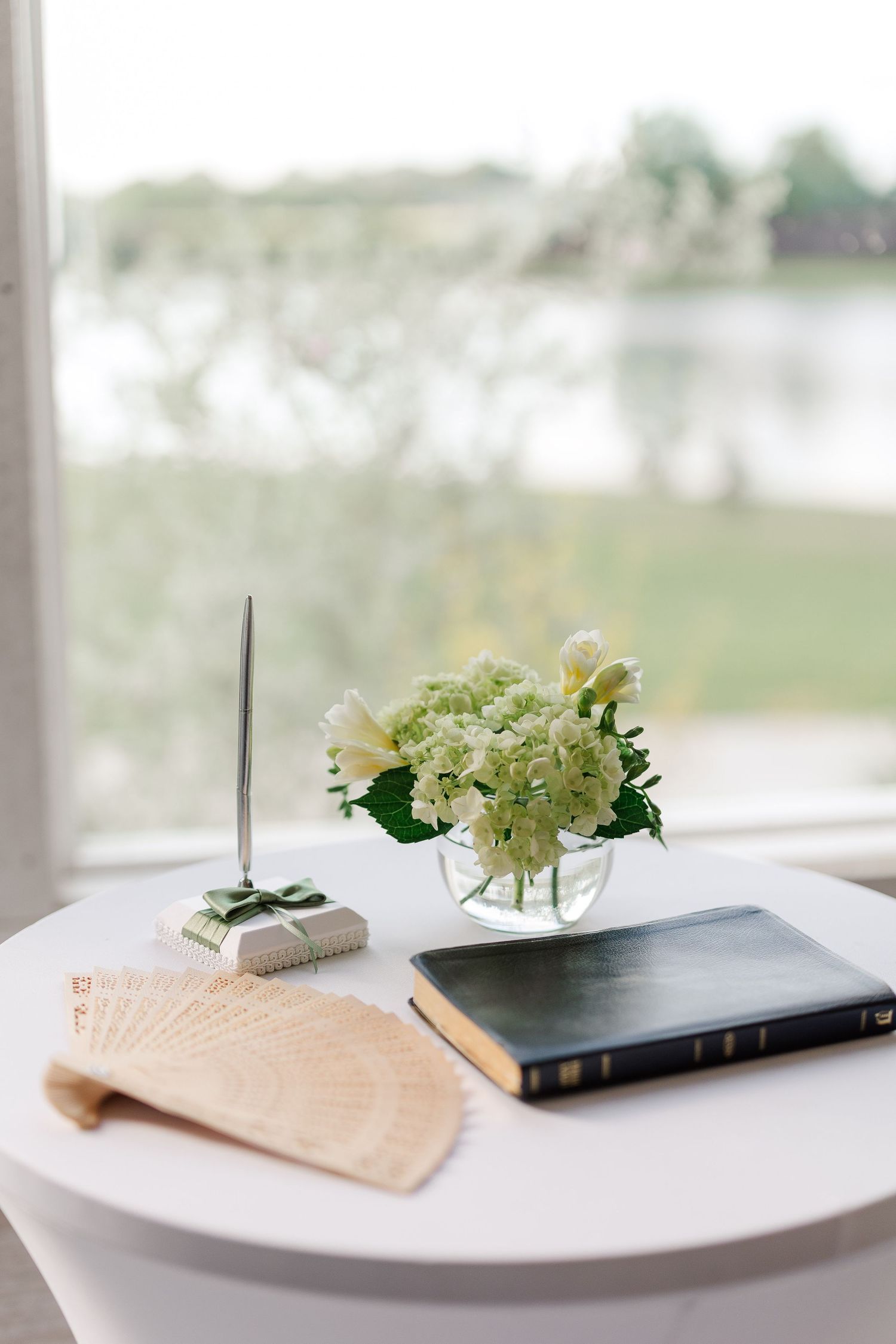 Elegant wedding guest book table with white hydrangeas, pen holder, wooden fan, and Bible by lakeside window.