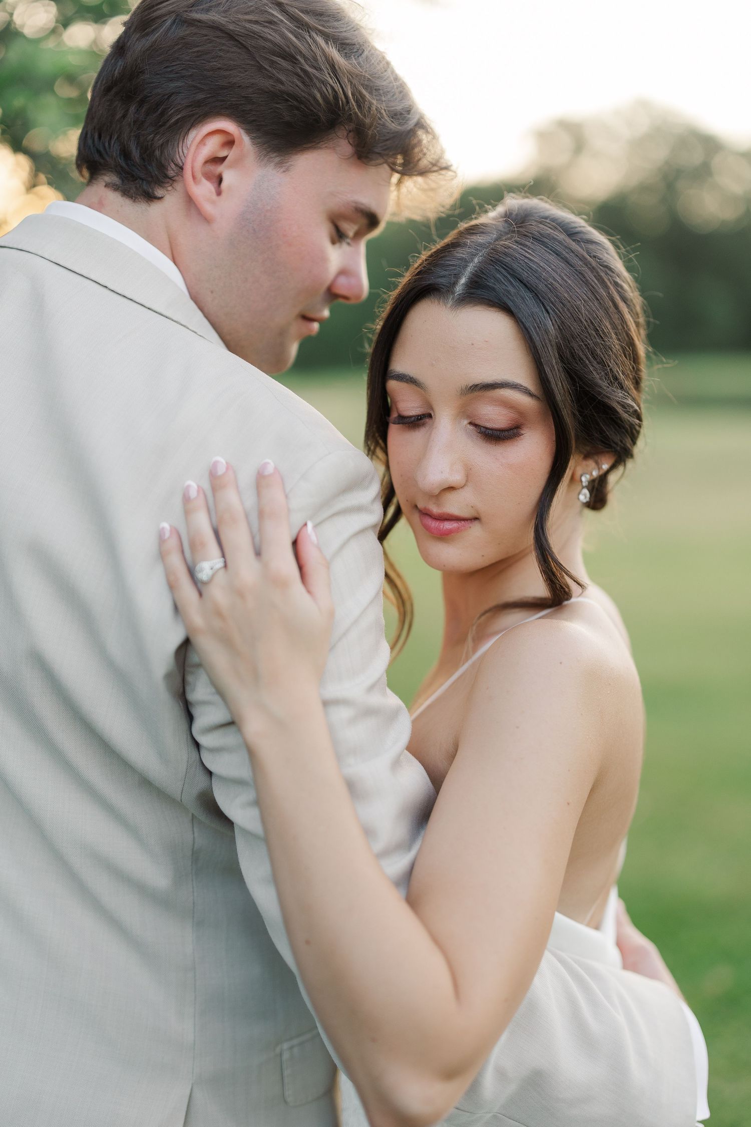 Bride in white gown embracing groom in beige suit during golden hour at an outdoor wedding.