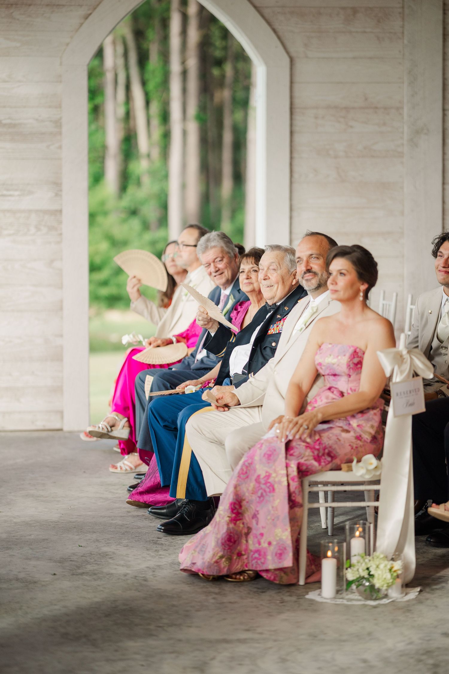 Wedding guests seated in white chapel ceremony space, fanning themselves, wearing colorful formal attire.