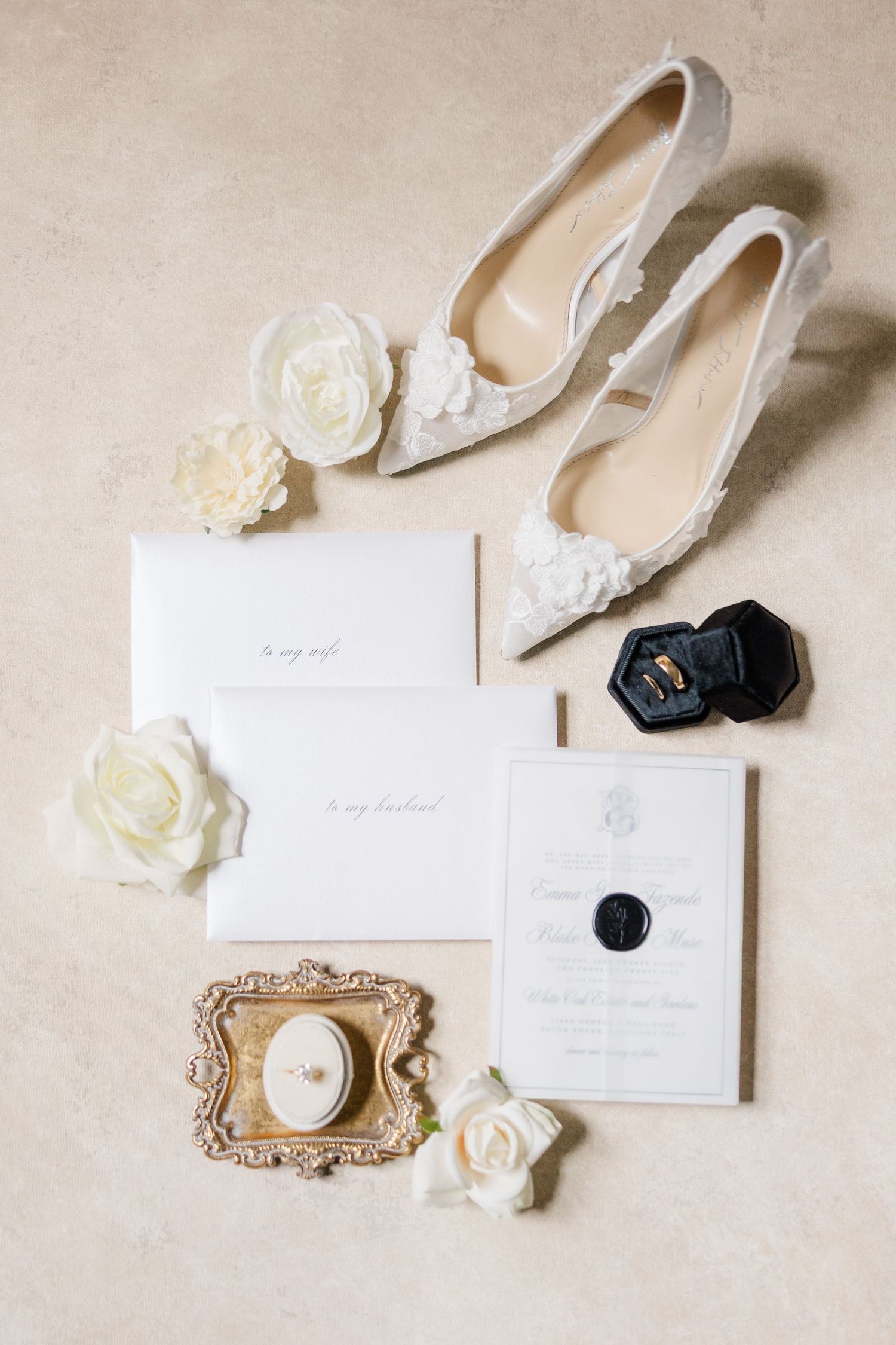 Elegant wedding accessories including white invitations, silver heels, delicate roses, and vintage gold tray arranged on beige background.