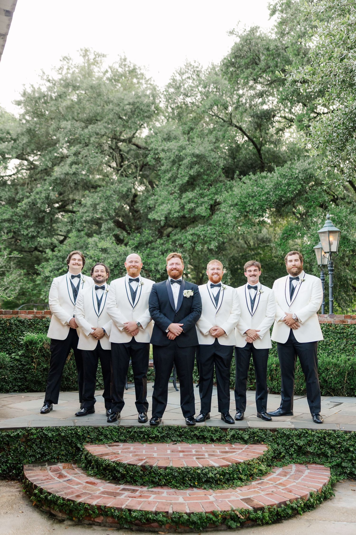 Groomsmen in white tuxedo jackets and black bowties pose on brick steps in a garden setting.